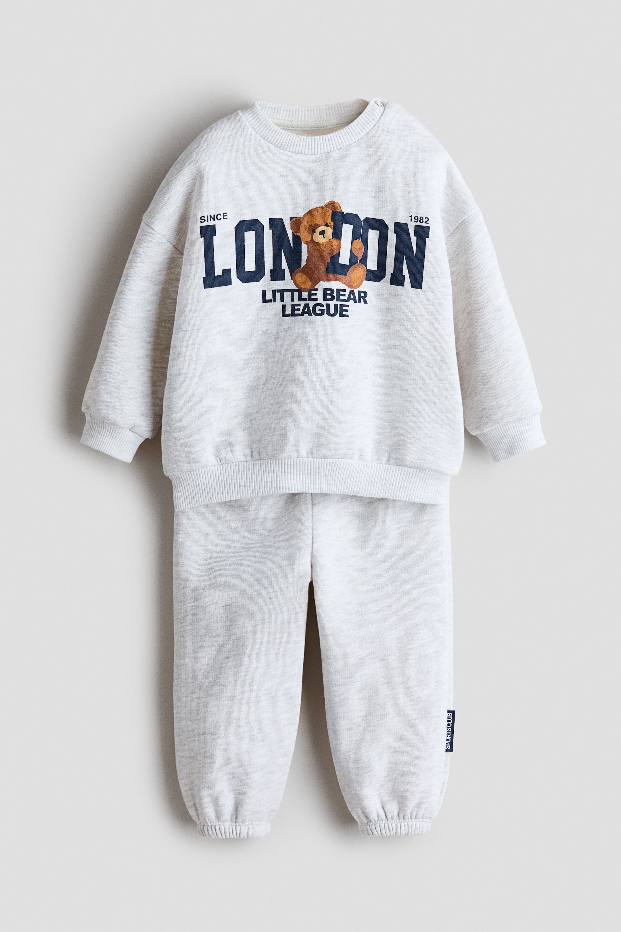 Visa större bild: Detta tvådelade sweatshirtset med tryck i ljusgrå melering visas framifrån och består av en långärmad sweatshirt med rund hals och matchande mjukisbyxor. Sweatshirten har ett marinblått grafiskt tryck med ordet "LONDON" integrerat med en nallebjörnsillustration på bröstet, medan mjukisbyxorna har resår vid anklarna som är samlade.