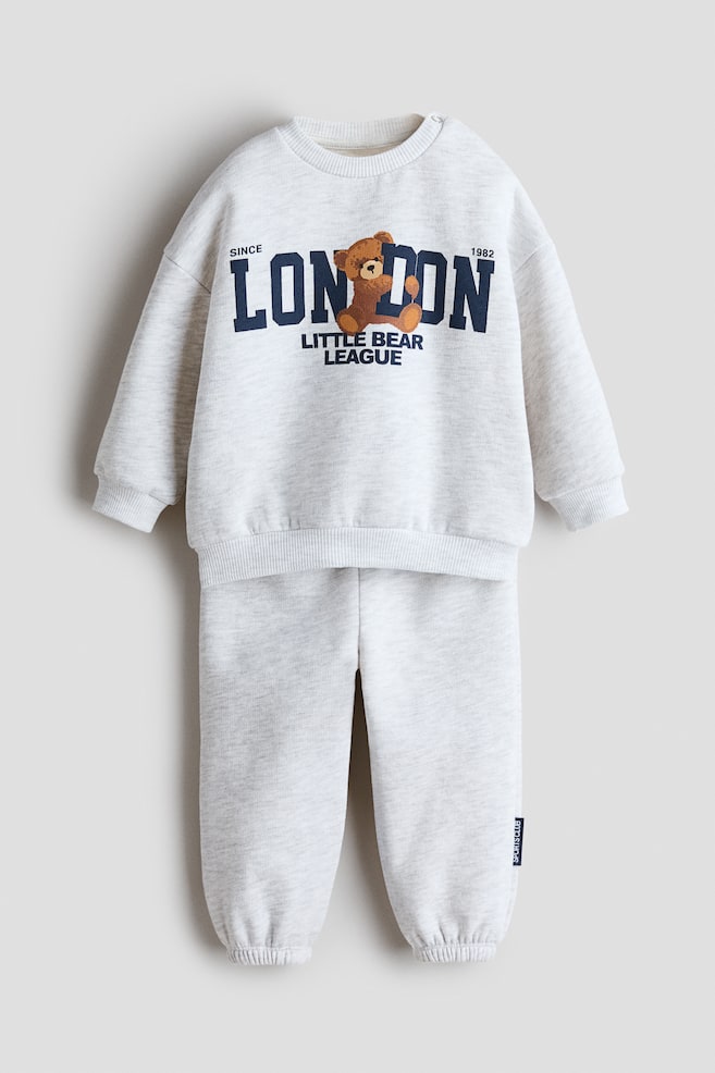 Dieses zweiteilige, bedruckte Sweatshirt-Set in hellgrauem Heather-Look ist von vorne zu sehen und prÃ¤sentiert ein langÃ¤rmliges Sweatshirt mit Rundhalsausschnitt sowie die dazu passenden Sweatpants. Auf der Brust zeigt das Sweatshirt einen marineblauen Grafikdruck des Wortes âLONDONâ, kombiniert mit einer TeddybÃ¤r-Illustration, wÃ¤hrend die Sweatpants mit gerippten, elastischen BÃ¼ndchen an den KnÃ¶cheln ausgestattet sind. - 1