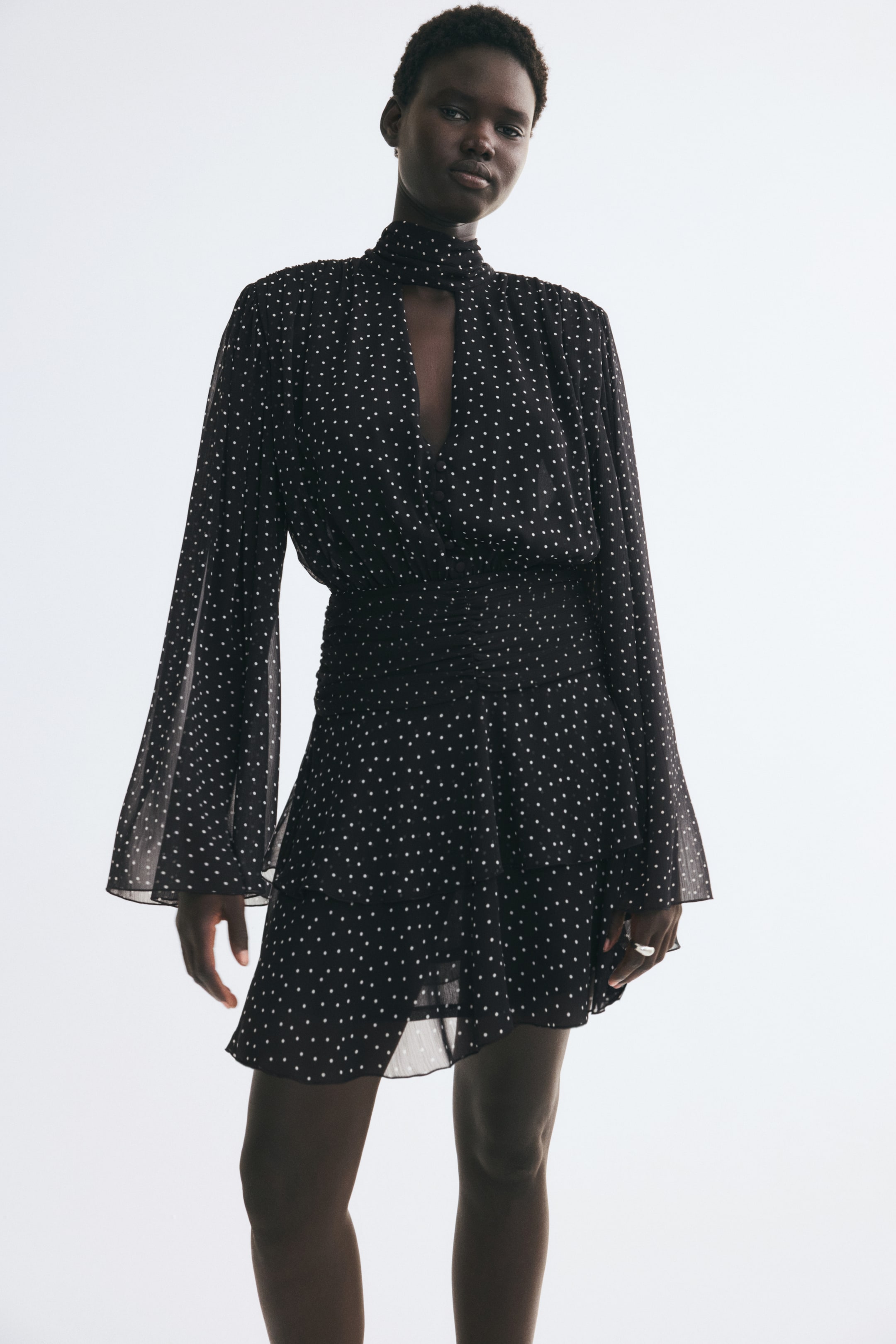 View larger image: The model is wearing a black-and-white polka-dot mini dress in crinkled chiffon. The dress features a high stand collar with a V-shaped front opening, long flared trumpet sleeves, a wide pleated waistband, and a tiered, gently flared skirt.