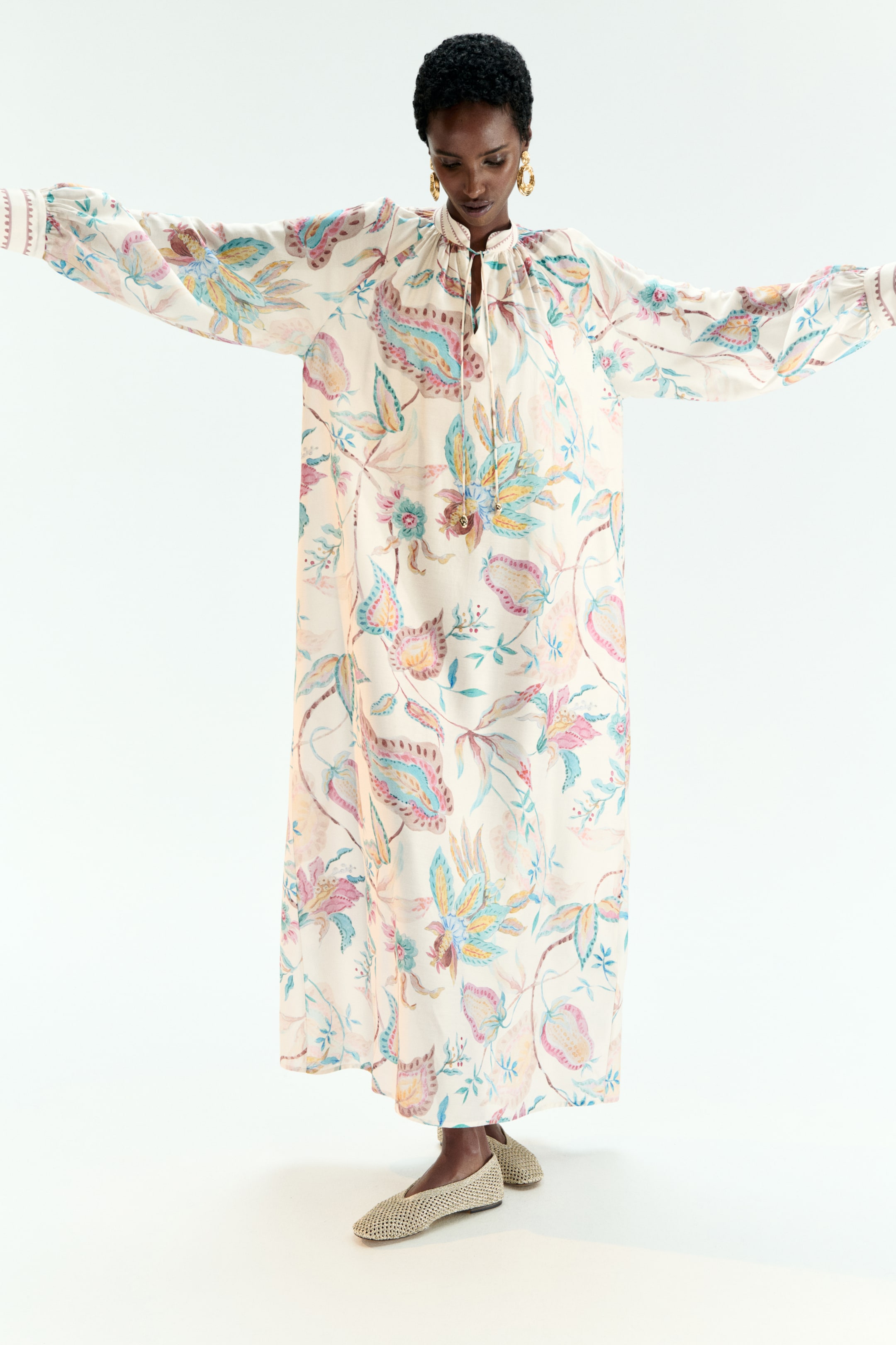 Visa större bild: En krämfärgad maxiklänning med ett färgglatt blommigt och paisley-mönster, med en ståkrage och V-ringning och knytning. Den har långa, vida ärmar som slutar i broderade manschetter och en flödande, avslappnad silhuett.