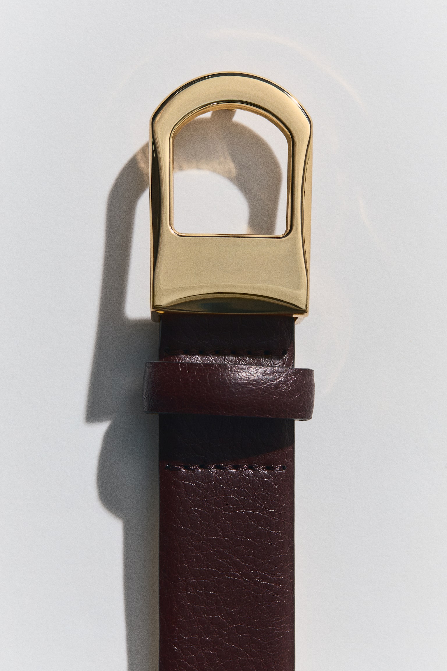 A dark brown leather belt lies flat and vertically oriented, showcasing a fine-grained texture. It features a polished gold-toned stirrup-shaped buckle, a matching leather strap keeper, and several adjustment holes along the strap. - 1