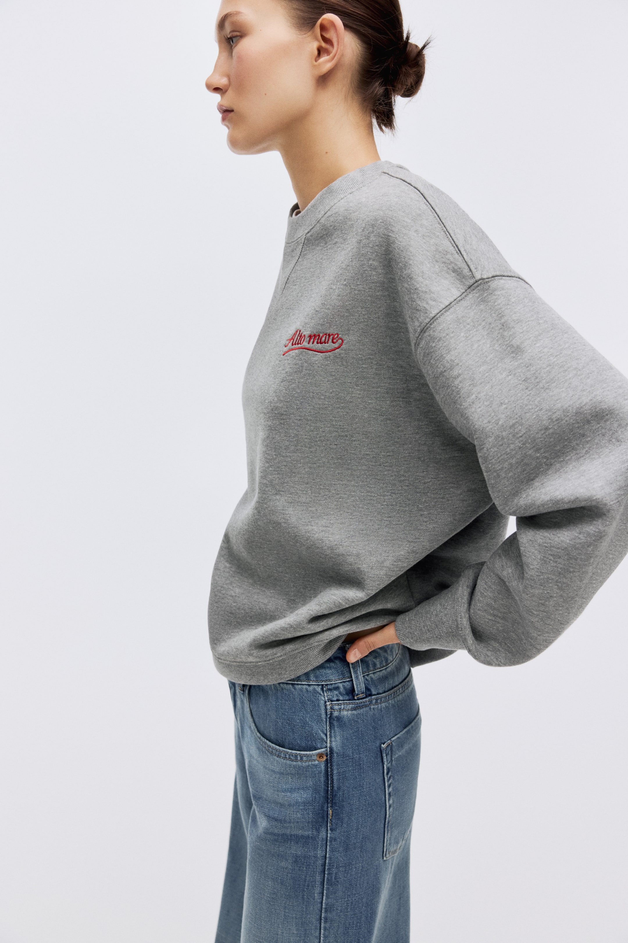 View larger image: A grey crewneck sweatshirt, shown from a side-front view, features red "Alto mare" embroidery on the left chest, with a relaxed fit, long sleeves, and a ribbed hem that sits over blue jeans.