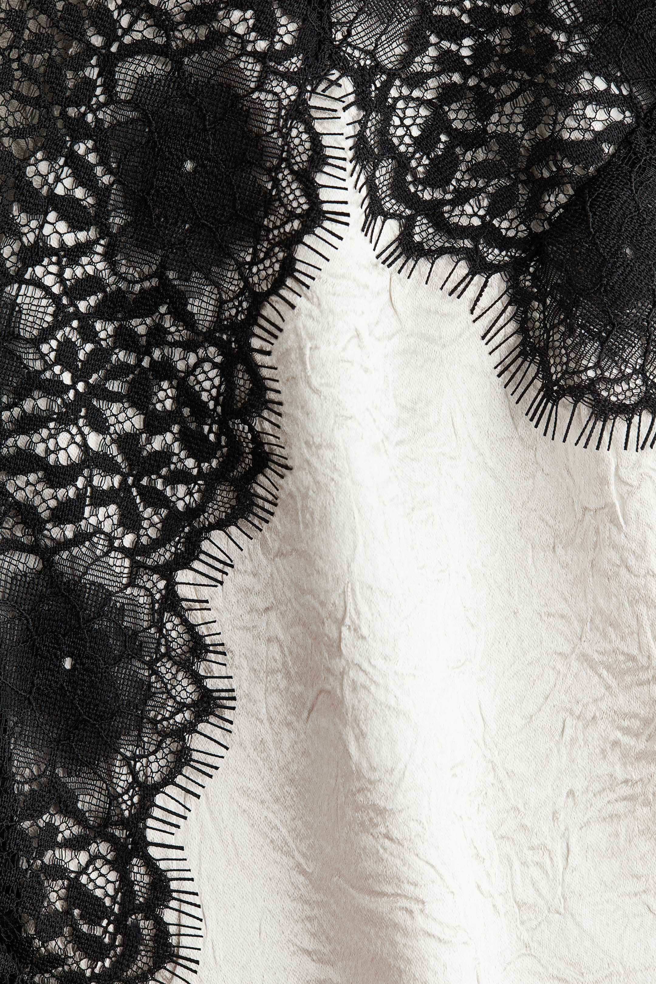 Zobraziť väčší obrázok: Textilný výrobok obsahuje čiernu čipku so zložitým vzorom a zvlneným okrajom, detailne spracovanú s krátkymi, výraznými niťami. Táto čipka ohraničuje svetlú, pokrčenú tkaninu.