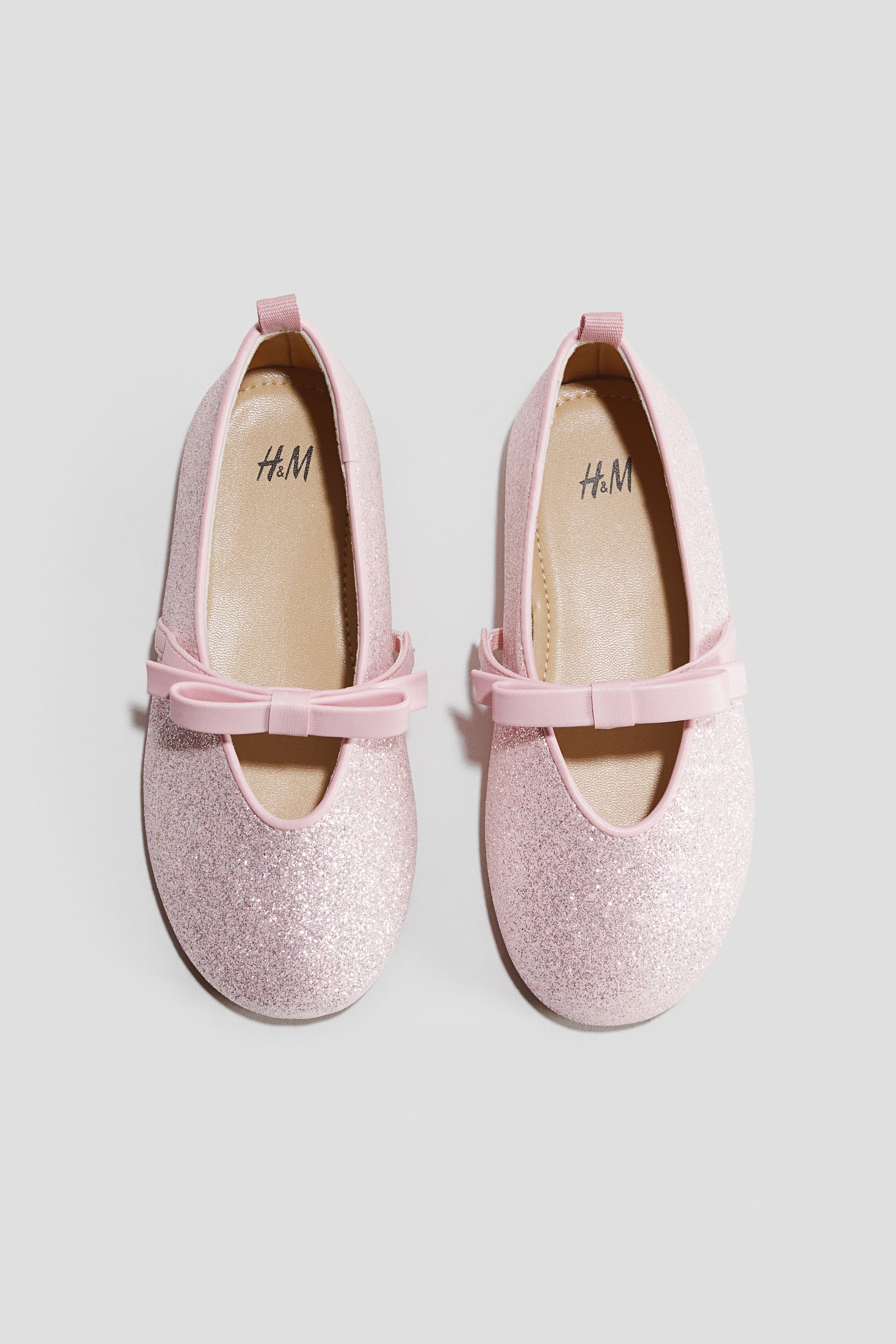 View larger image: A pair of pink glitter ballet flats features a round toe and an elastic strap with a pink bow across the vamp. Each shoe also displays a small pink pull tab at the heel and the H&M logo on the beige insole.