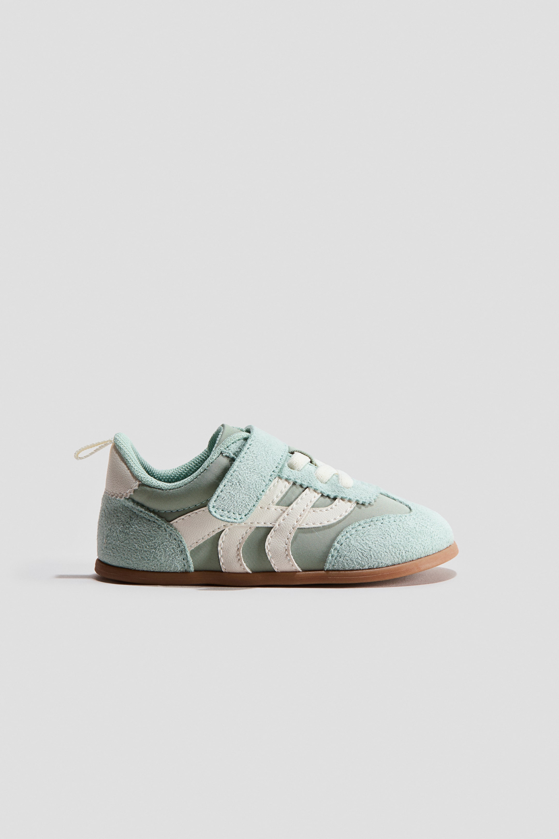 View larger image: Trainer. A light green trainer is depicted from the side, showcasing light green suede-like panels on the toe and heel, complemented by a single velcro strap closure. White stripes accentuate the midfoot, while a light brown sole is visible underneath.