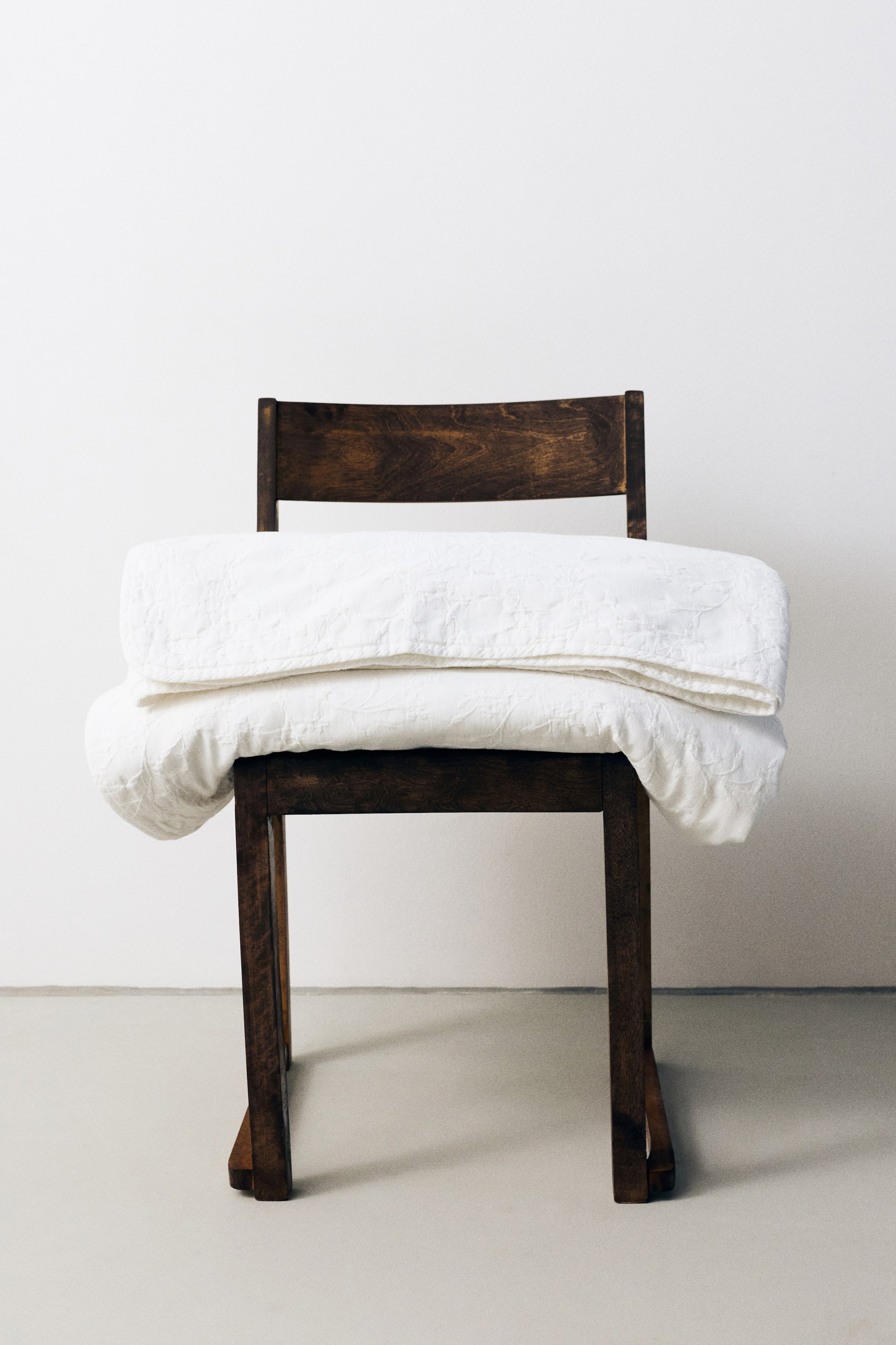 View larger image: A white, lightly padded cotton bedspread featuring a textured floral jacquard weave is draped in a folded style over the seat of a dark wooden chair, displaying its soft structure and subtle pattern.