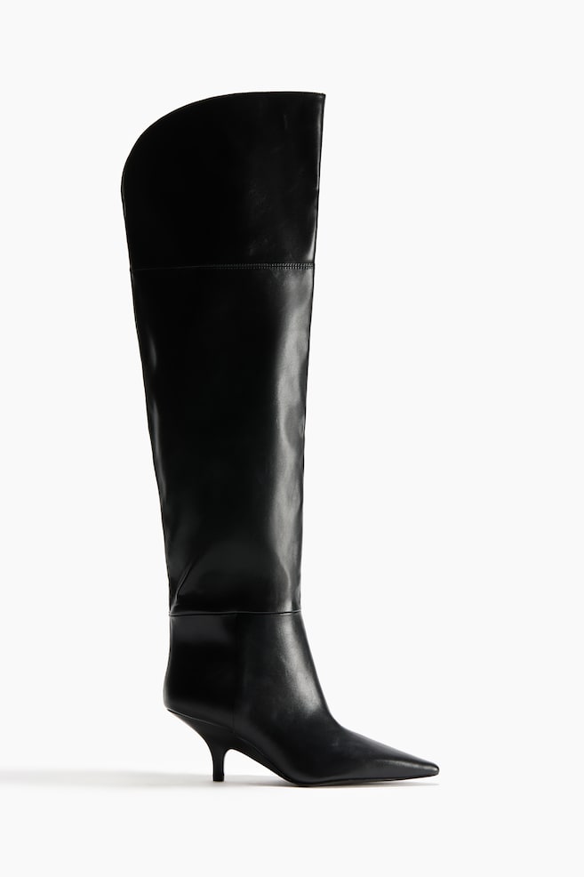 A black leather over-the-knee boot is shown in profile facing right, featuring a pointed toe, a low kitten heel, and a smooth leather finish. The boot features a tall, structured shaft with an asymmetrical curved topline that rises higher at the front, accented by minimal horizontal seam detailing at the ankle and upper leg. - 1