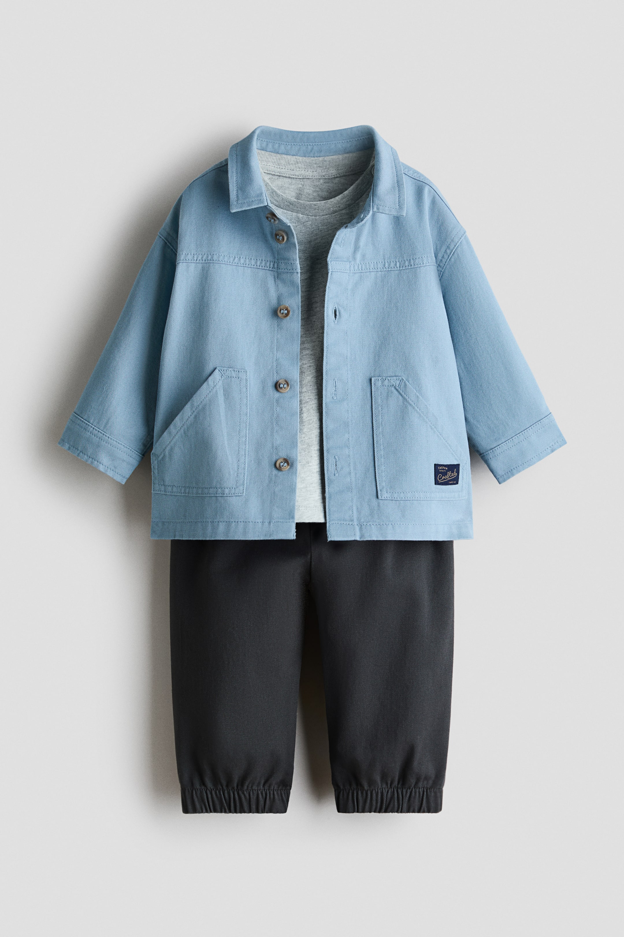 View larger image: 3-piece cotton set, FRONT. A light-blue collared jacket shows front button closures and two large patch pockets, layered over a light-grey crew-neck T-shirt. Dark-grey jogger-style trousers complete the set with elasticated cuffs.