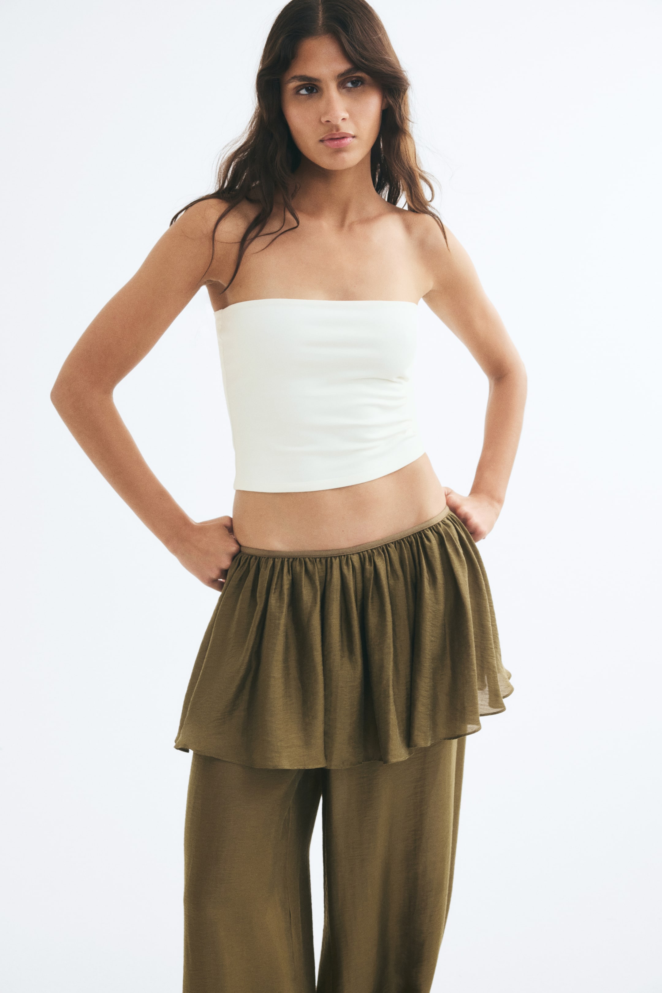 View larger image: A white, fitted bandeau top with a strapless design and a straight-cut hem is worn by the model. The top is paired with olive green, wide-leg trousers featuring a gathered, ruffled waist.