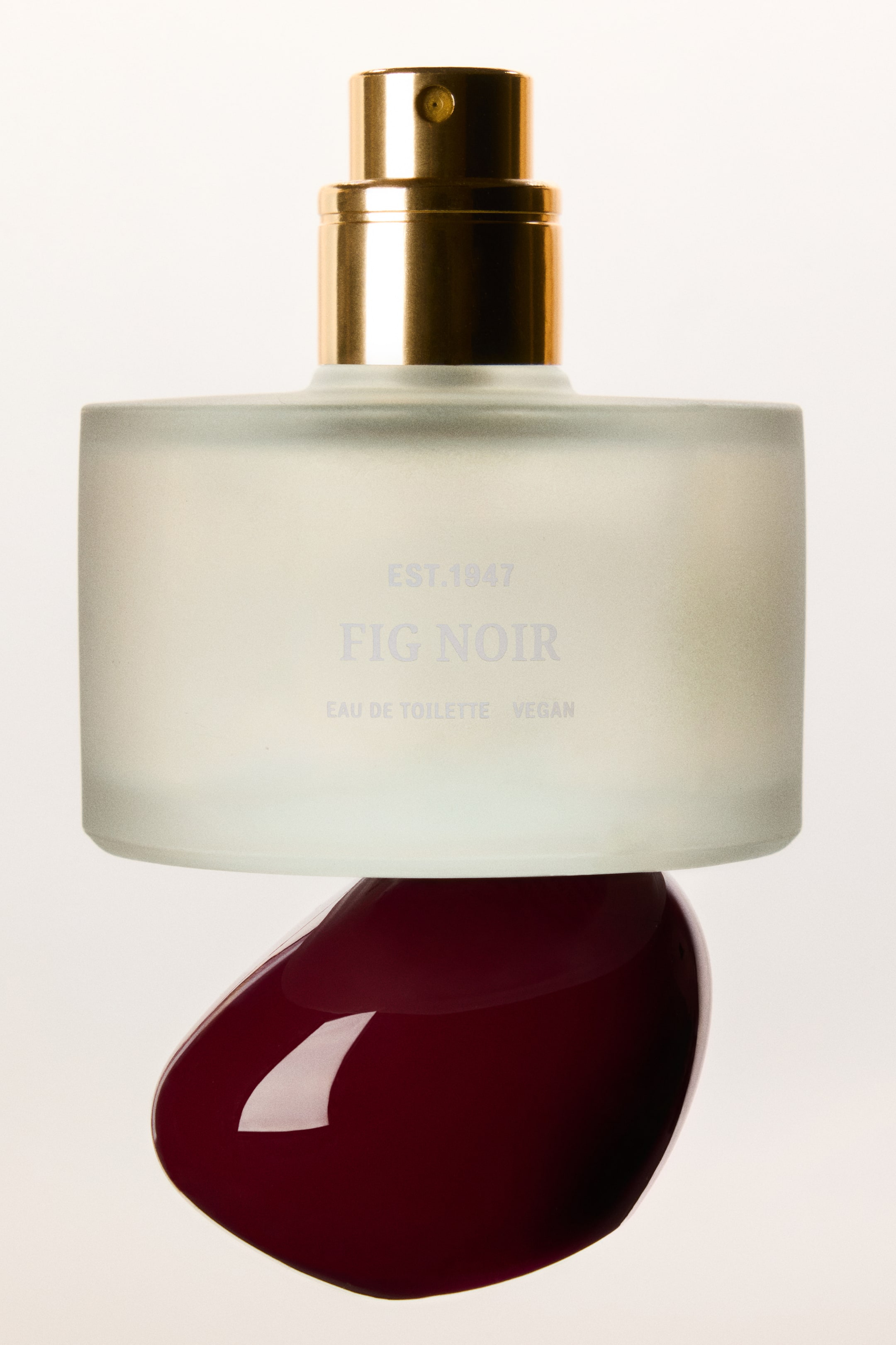 Zobraziť väčší obrázok: Fig Noir Eau De Toilette je prezentovaná ako široký valcovitý matný sklenený flakón so zlatisto tónovanou rozprašovacou tryskou, ktorý je vyvážený na hladkom, lesklom tmavočervenom organicky tvarovanom kameni. Flakón otočený spredu zobrazuje svetlosivý potlačený text s nápisom „EST.1947 FIG NOIR EAU DE TOILETTE VEGAN“.