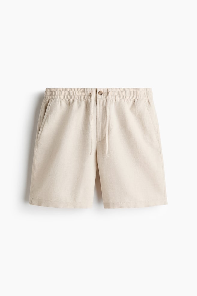 Light beige shorts are presenting a front view, laid flat, featuring an elasticated waistband with a drawstring and a button closure. Side seam pockets are visible, and the fabric is showing a textured cotton and linen weave. - 1