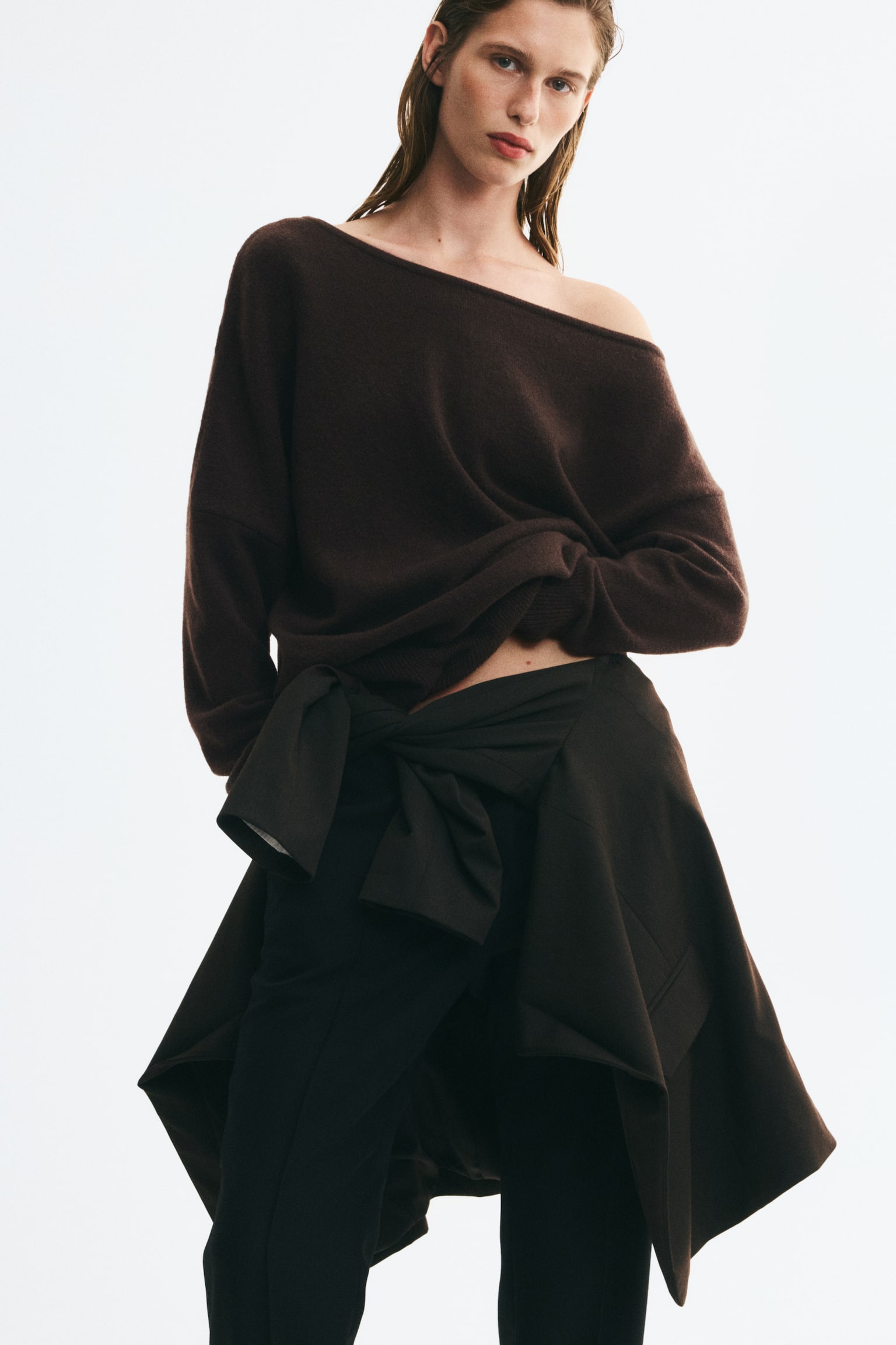 A dark brown, fine-knit sweater featuring an asymmetrical one-shoulder neckline and dropped shoulders is being worn by the model. The sweater is paired with black trousers and a dark jacket tied around the waist, with the model holding the hem slightly gathered on one side. - 1