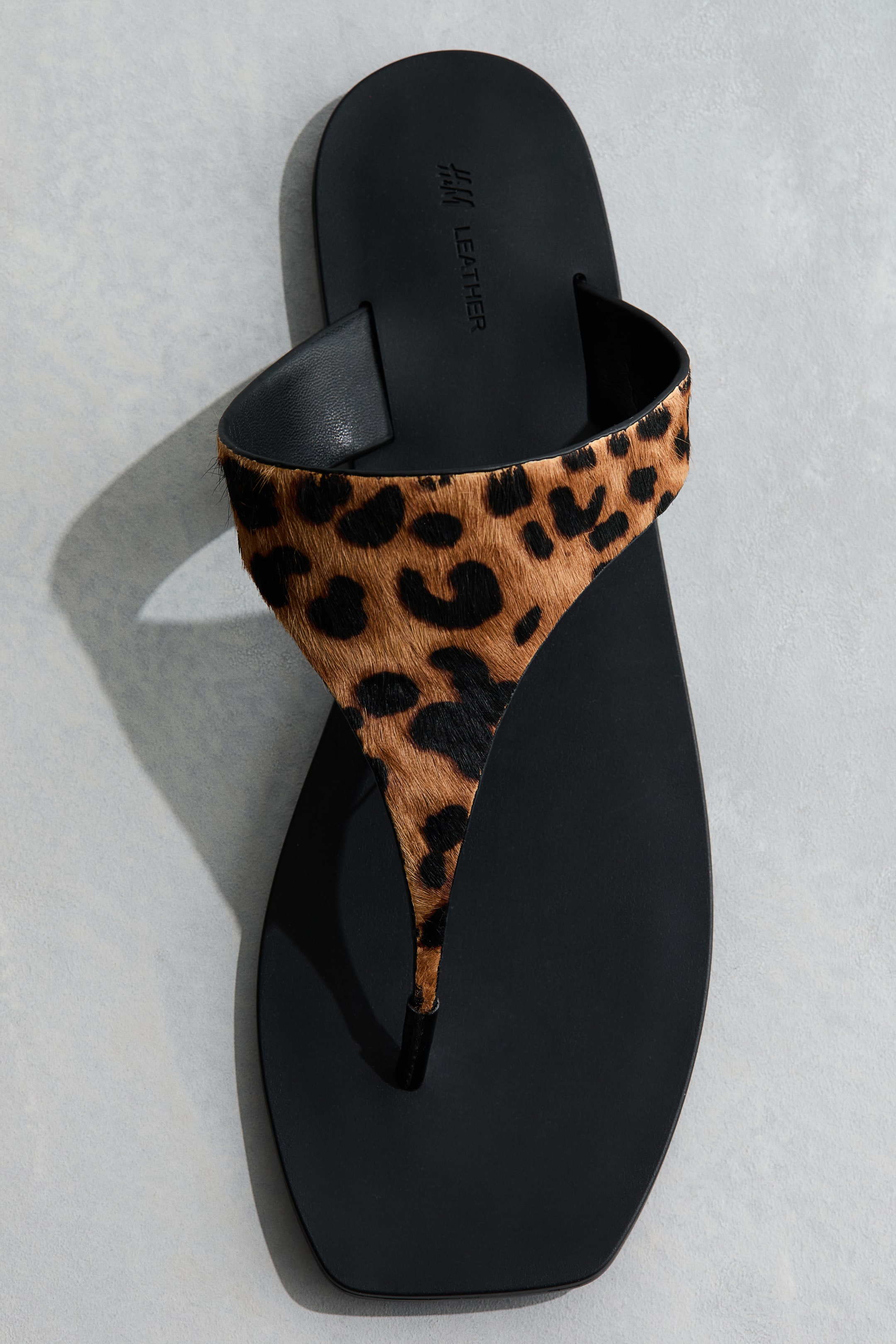 View larger image: A single black thong sandal showcases a wide strap across the mid-foot, adorned with a leopard print pattern in brown and black. The sleek black footbed displays "H&M LEATHER" embossed near the top.