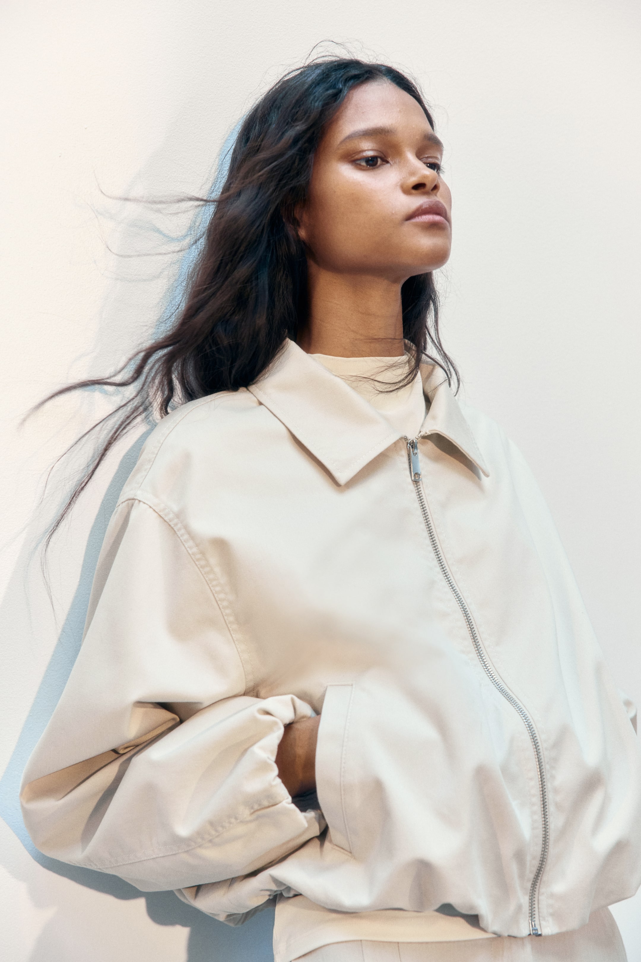 Grotere afbeelding bekijken: Een oversized off-white twill jasje met een kraag en een gedeeltelijk ritssluiting wordt gedragen door het model. Het model steekt één hand in een paspel zijzak, terwijl de elastische boorden en zoom van het jasje een volumineus silhouet creëren over een zichtbaar lichtgekleurde top.