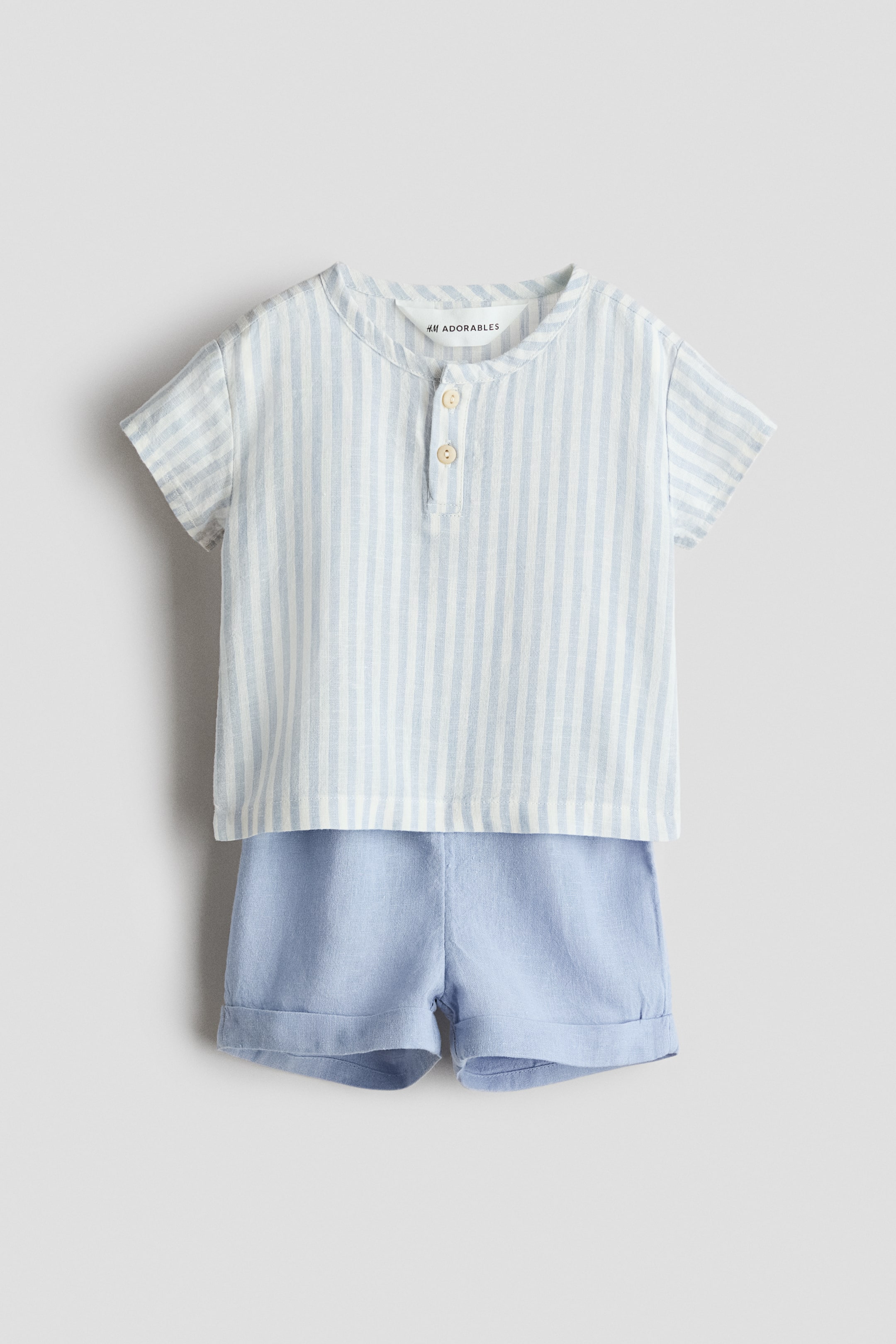 View larger image: A baby’s two-piece set, shown from the front, comprises a short-sleeved shirt with light blue and white vertical stripes, a round neckline and a two-button placket, paired with light blue shorts featuring rolled cuffs.