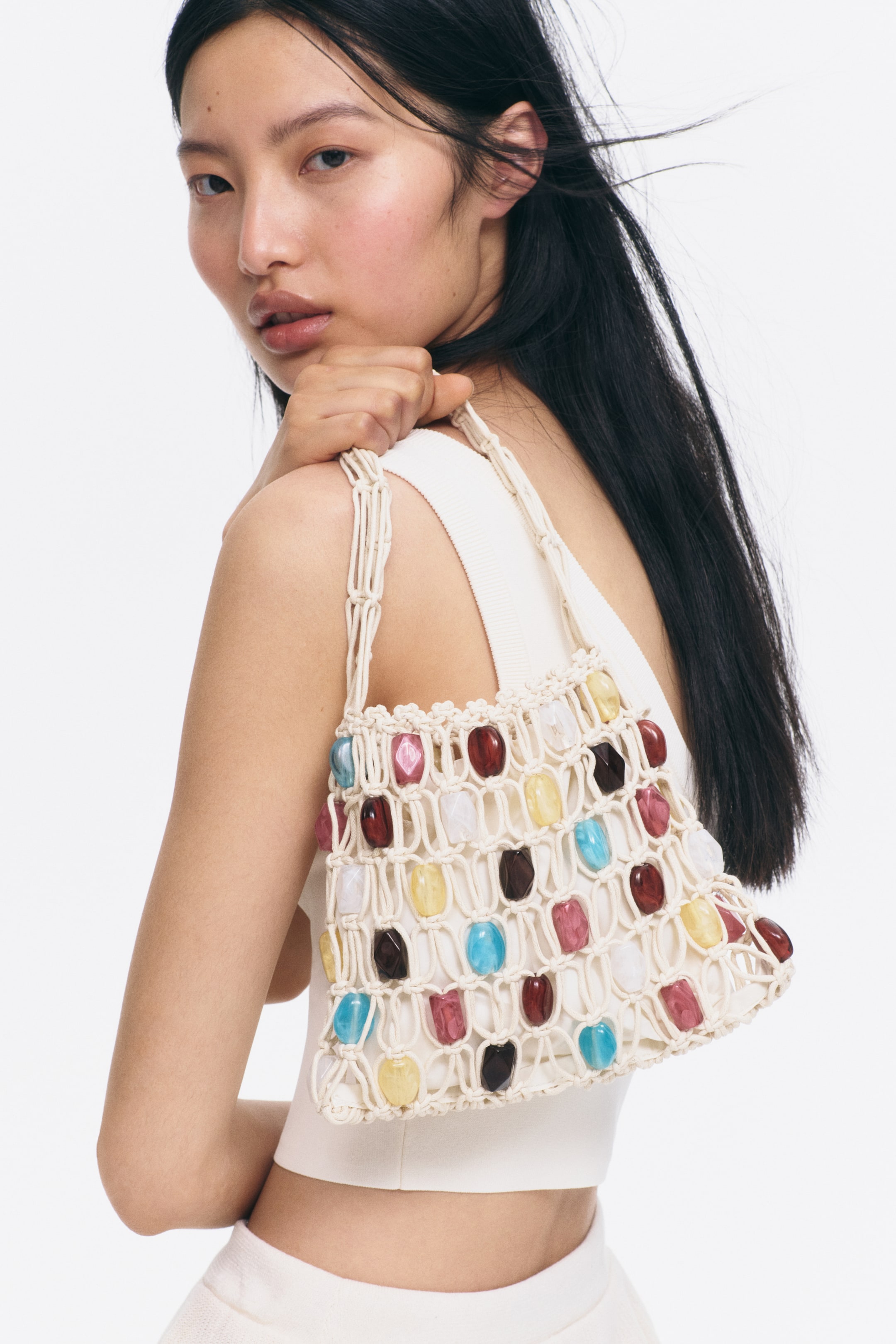 Agrandir l'image: Un sac à bandoulière en macramé de couleur crème est porté sur une épaule, orné de perles oblongues translucides multicolores intégrées dans son filet selon un motif en grille.