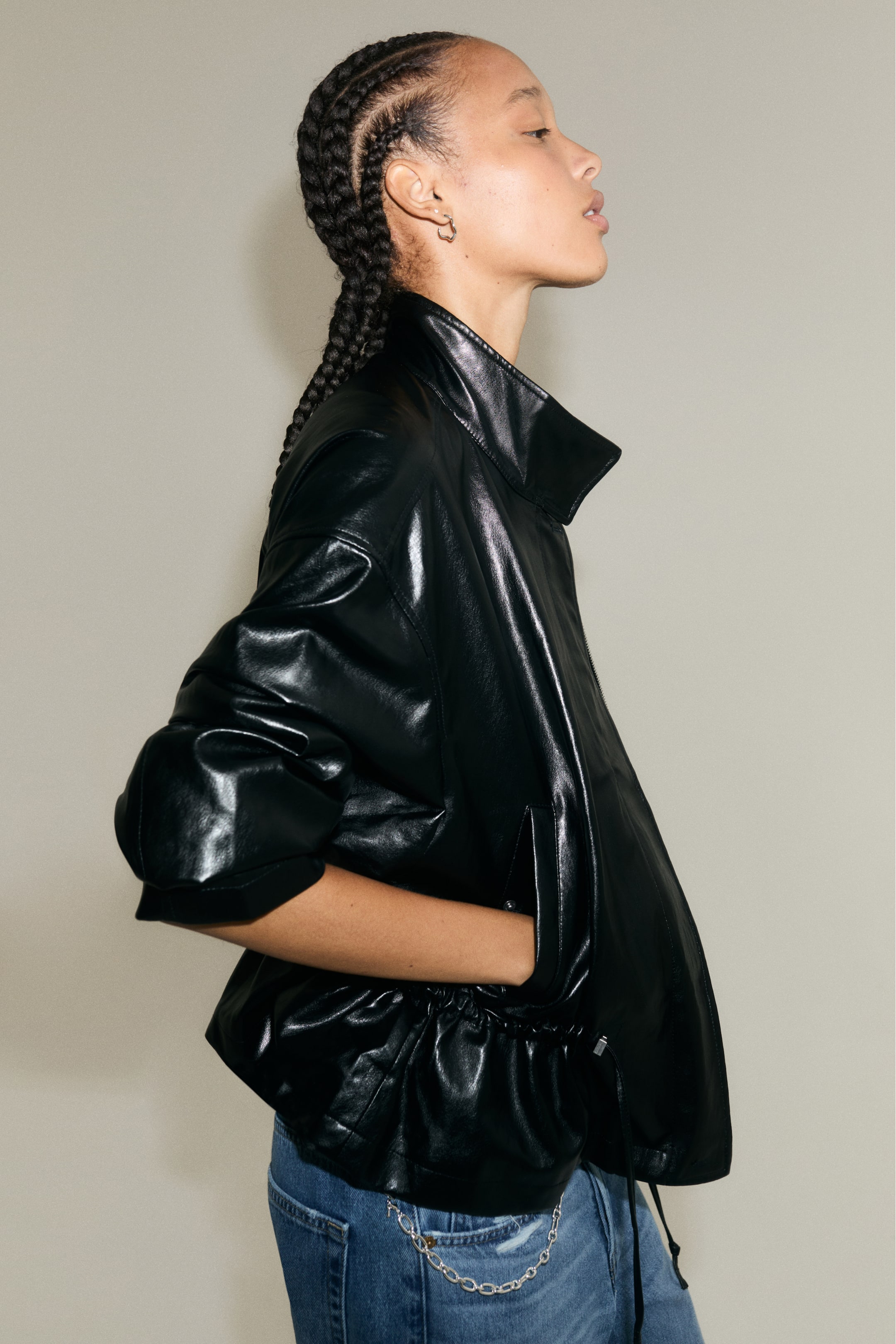 Größeres Bild anzeigen: Das Model trägt eine Oversize-Jacke aus glänzendem, schwarz beschichtetem Stoff und hat eine Hand in der Leistentasche vorne. Die Jacke verfügt über einen Stehkragen, überschnittene Schultern und einen gerafften Kordelzug in der Taille und ist mit blauen Jeans kombiniert.