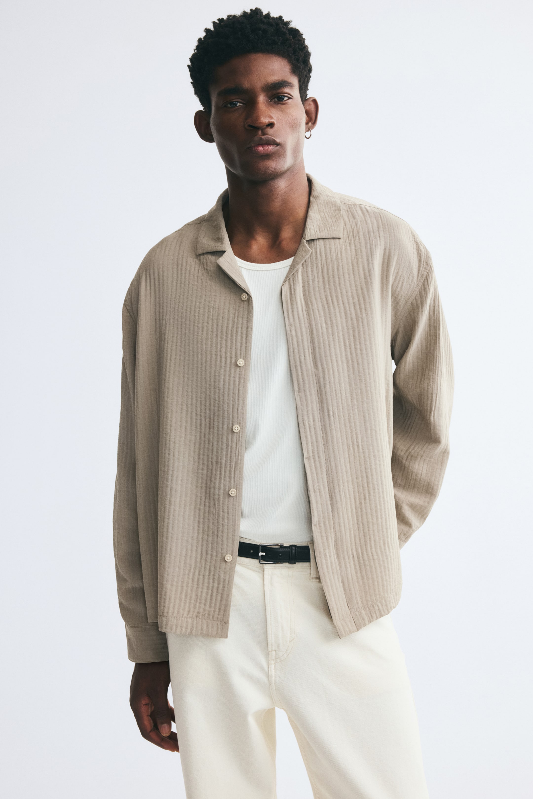 View larger image: A light beige, textured-weave long-sleeve shirt with a resort collar is worn unbuttoned by the model. The shirt is layered over a white ribbed tank top and paired with off-white trousers.