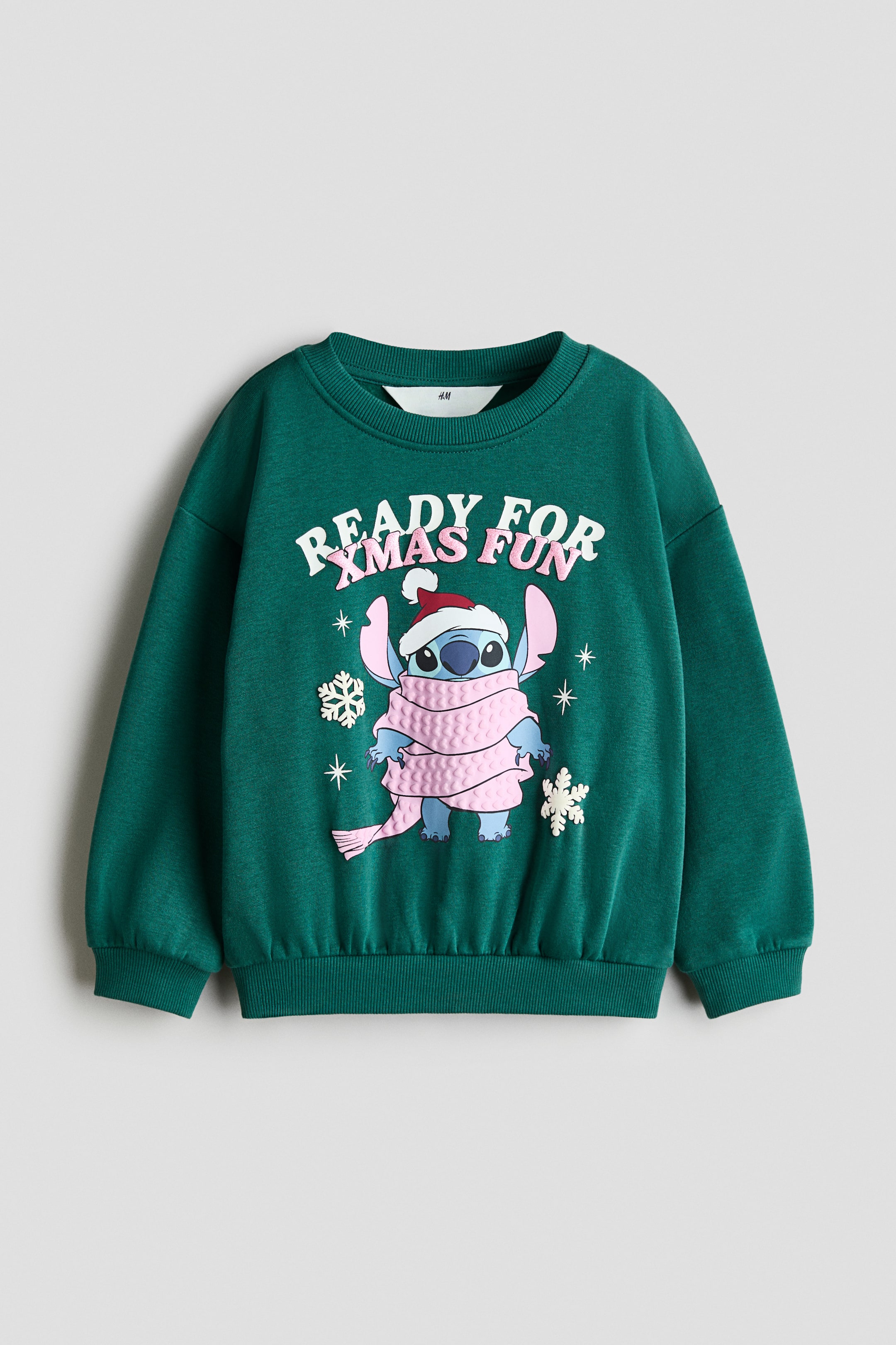 Voir une image plus grande: Un sweat-shirt vert à manches longues est présenté de face, avec un col rond côtelé, des épaules tombantes, ainsi qu’un ourlet et des poignets côtelés. L’avant affiche un graphique du personnage Stitch portant un bonnet de Père Noël et une écharpe rose texturée, avec le texte « READY FOR XMAS FUN » imprimé au-dessus en lettres blanches et roses, accompagné de petits détails de flocons de neige.