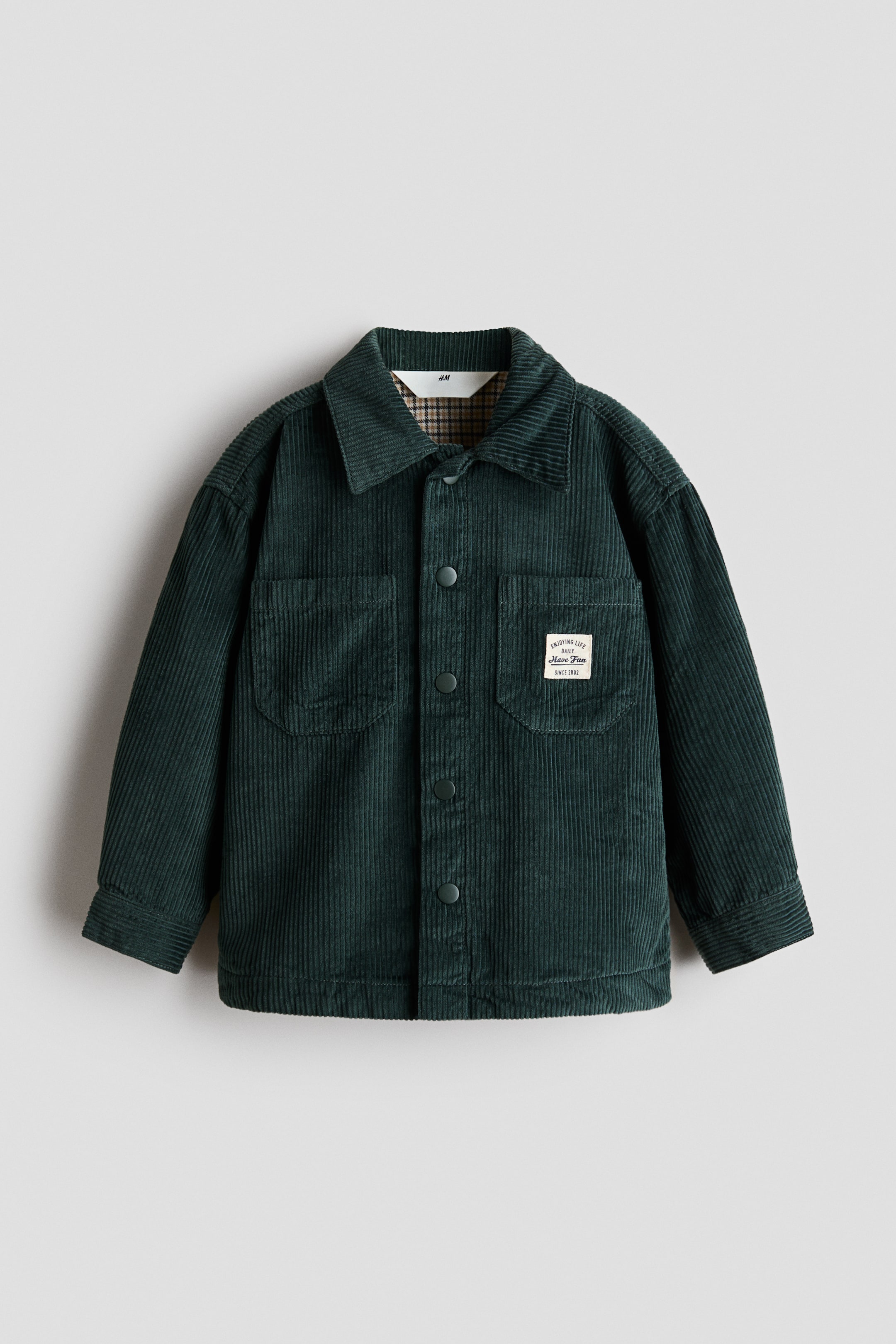 View larger image: A front-facing dark green corduroy shirt jacket features a point collar, a central snap-button closure, and two chest patch pockets. The left pocket has a small white rectangular patch, and a brown-and-white checkered lining is visible at the inner neckline.