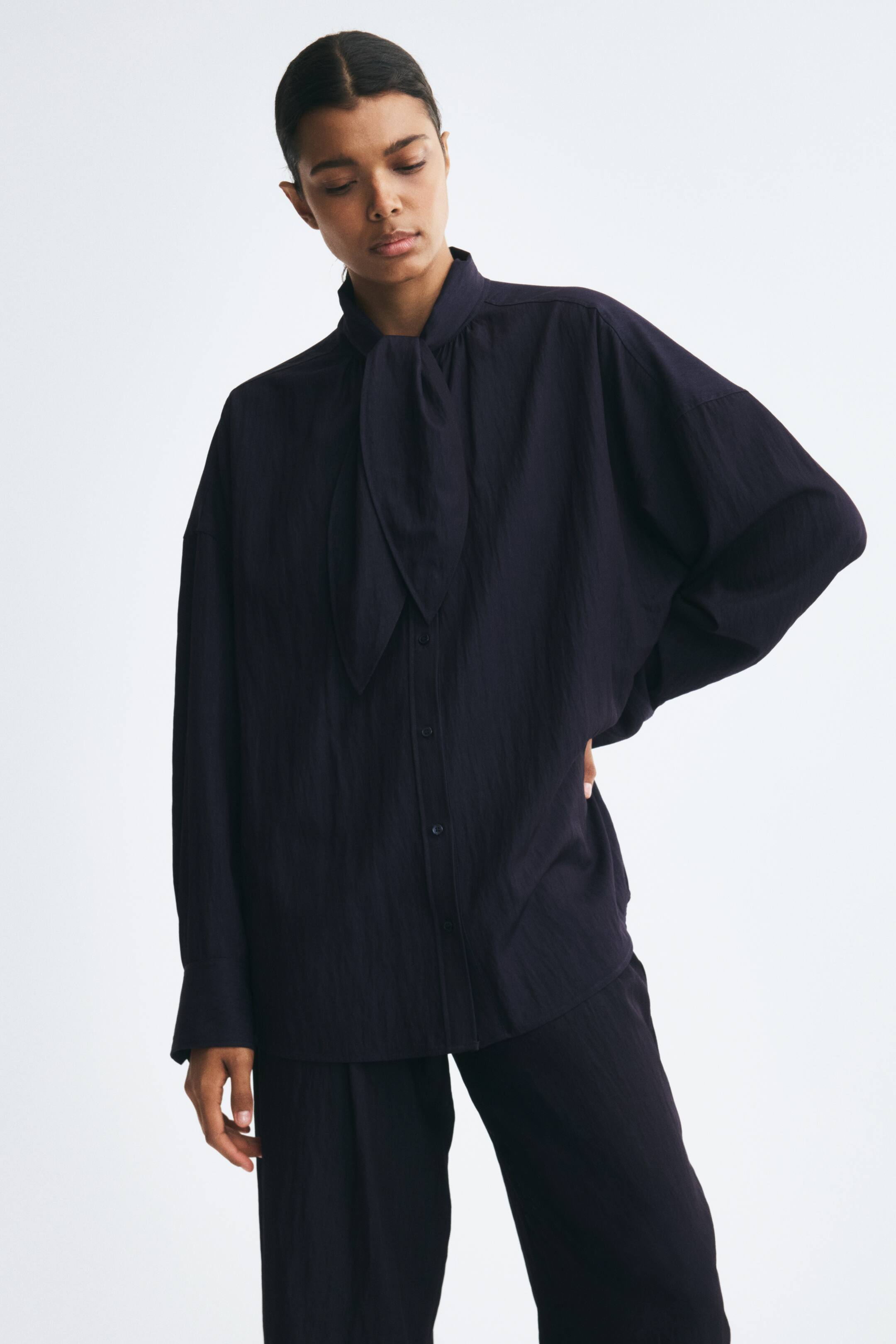 View larger image: A dark navy, textured button-up blouse with a tie-neck collar and long sleeves is worn by the model. The loose-fitting top is styled with matching trousers in the same fabric.
