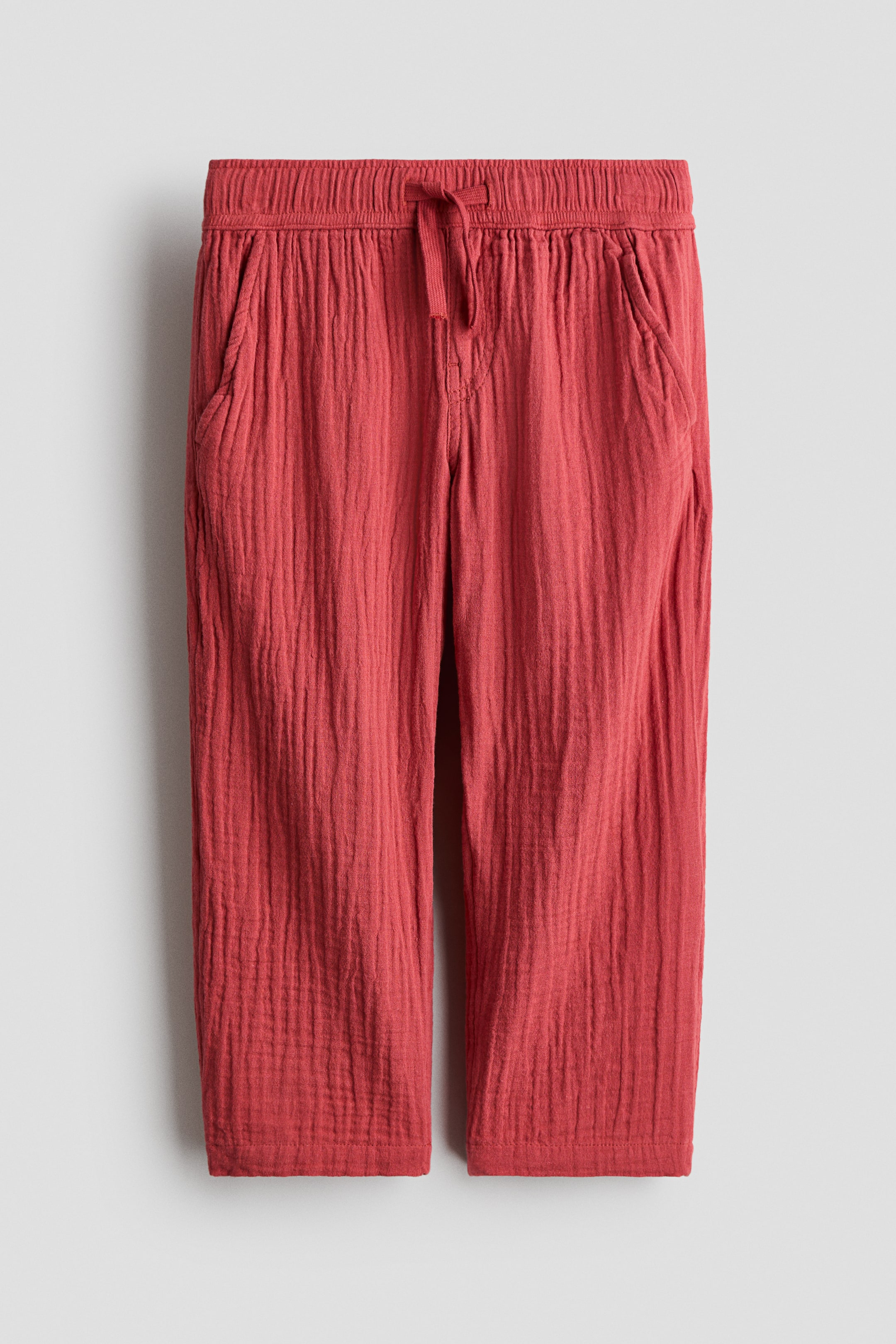 View larger image: Reddish-orange trousers are displayed from the front, showcasing a crinkled cotton muslin texture. They come with an elasticated, tied drawstring waist, a mock fly detail, diagonal side pockets, and straight legs.