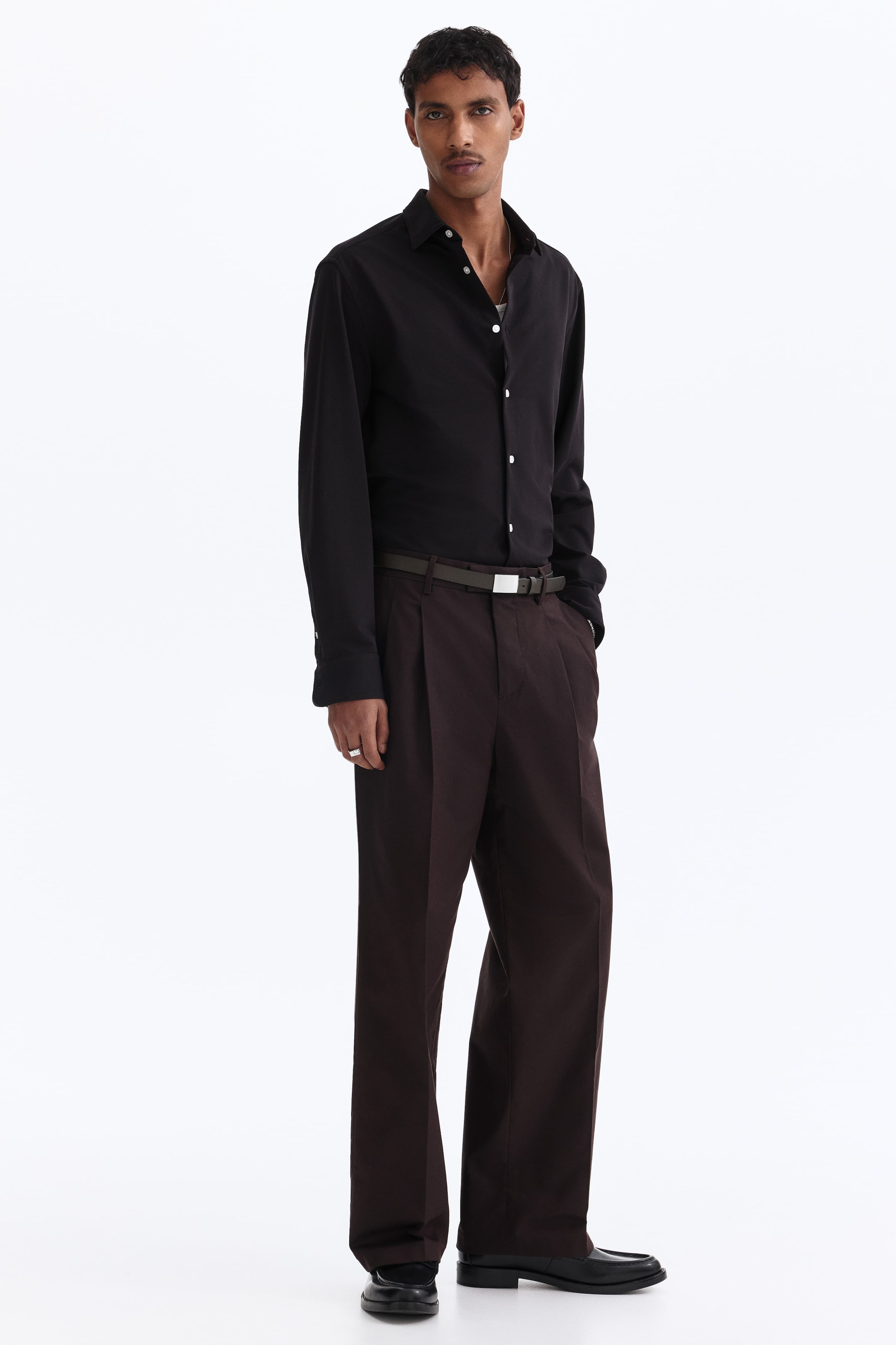 View larger image: A black collared shirt with white buttons along the front placket is tucked into dark brown wide-leg pleated trousers. The trousers are cinched with an olive-green belt featuring a silver-toned rectangular buckle.