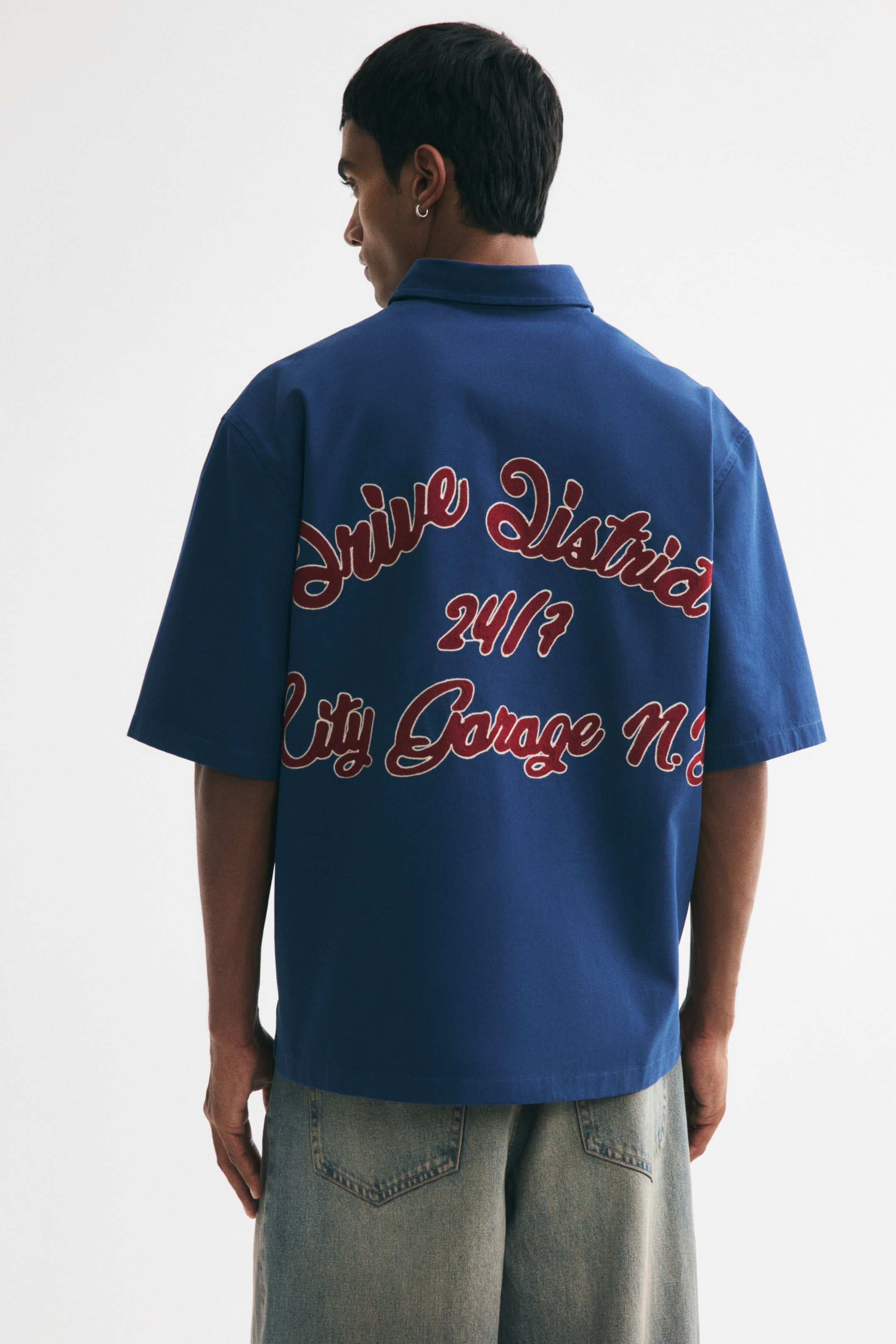 View larger image: The model is wearing a blue cotton twill shirt featuring a collar, short sleeves, a relaxed fit, and a straight-cut hem. On the back yoke of the shirt, there's a red and white embroidered motif that reads "Drive District 24/7 City Garage N.Y." The look is completed with light-wash jeans.
