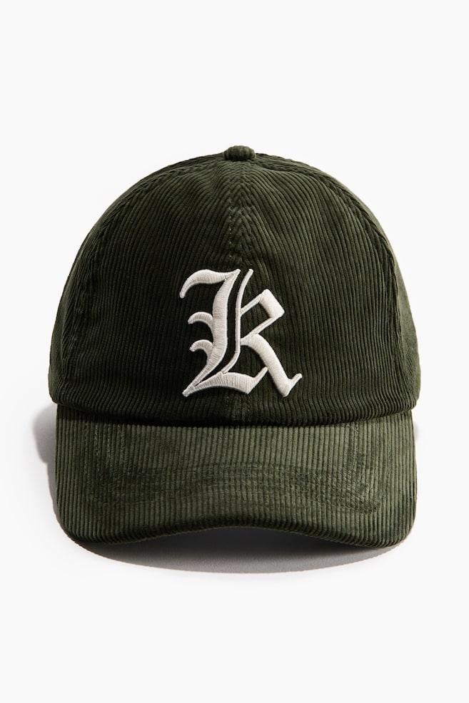 A dark green corduroy cap is presenting its front, showcasing a white embroidered stylized R motif on the crown. - 1