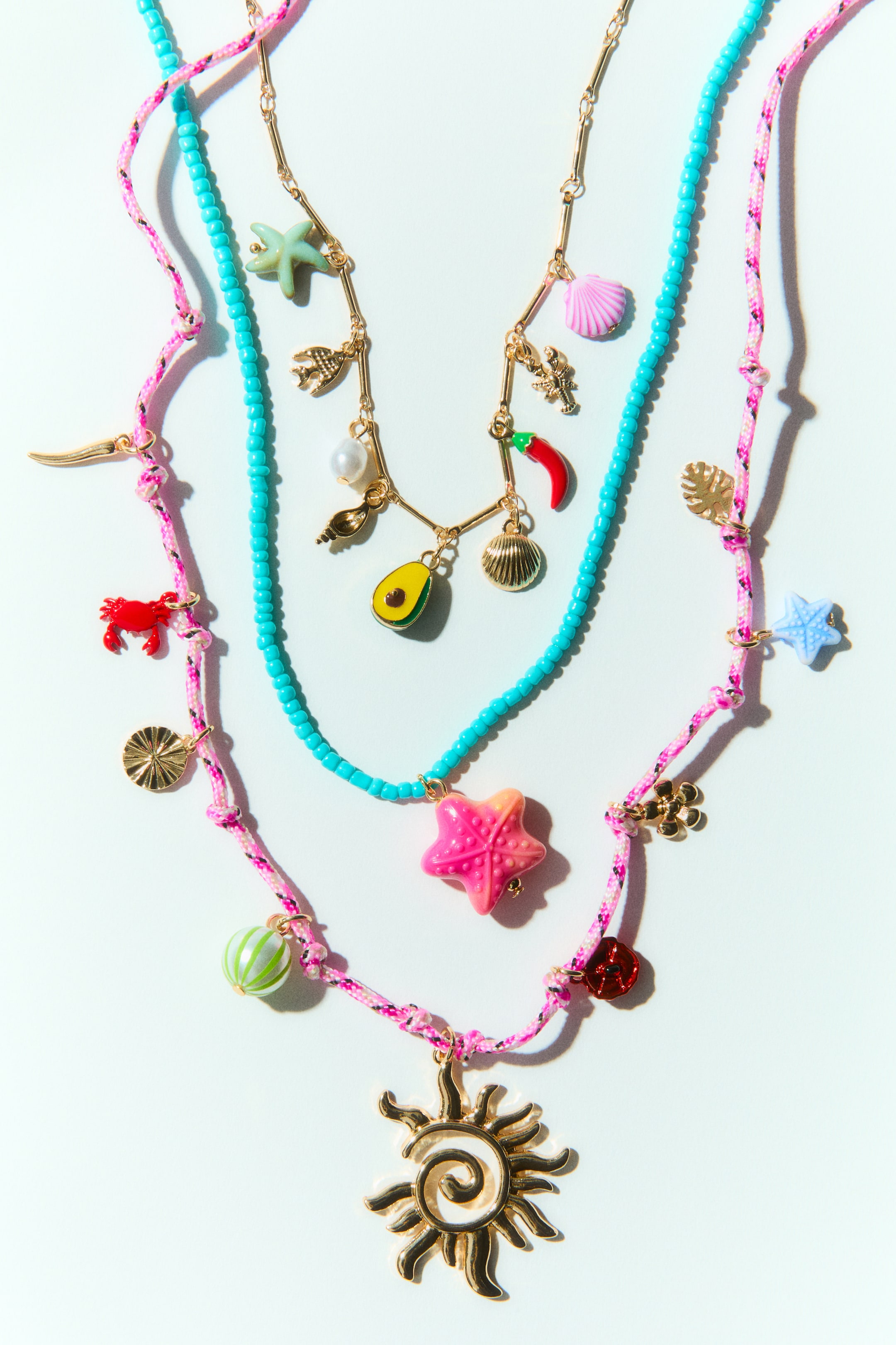 Grösseres Bild ansehen: Eine dreireihige Halskette wird von vorne präsentiert und zeigt ihr geschichtetes Design. Sie besteht aus einer goldfarbenen Metallgliederkette mit verschiedenen Anhängern, einem türkisfarbenen Perlenstrang mit einem großen rosa Sternanhänger und einem rosa geknüpften Kordelstrang, der eine Auswahl an Anhängern, darunter ein prominenter Sonnenanhänger, zur Schau stellt.