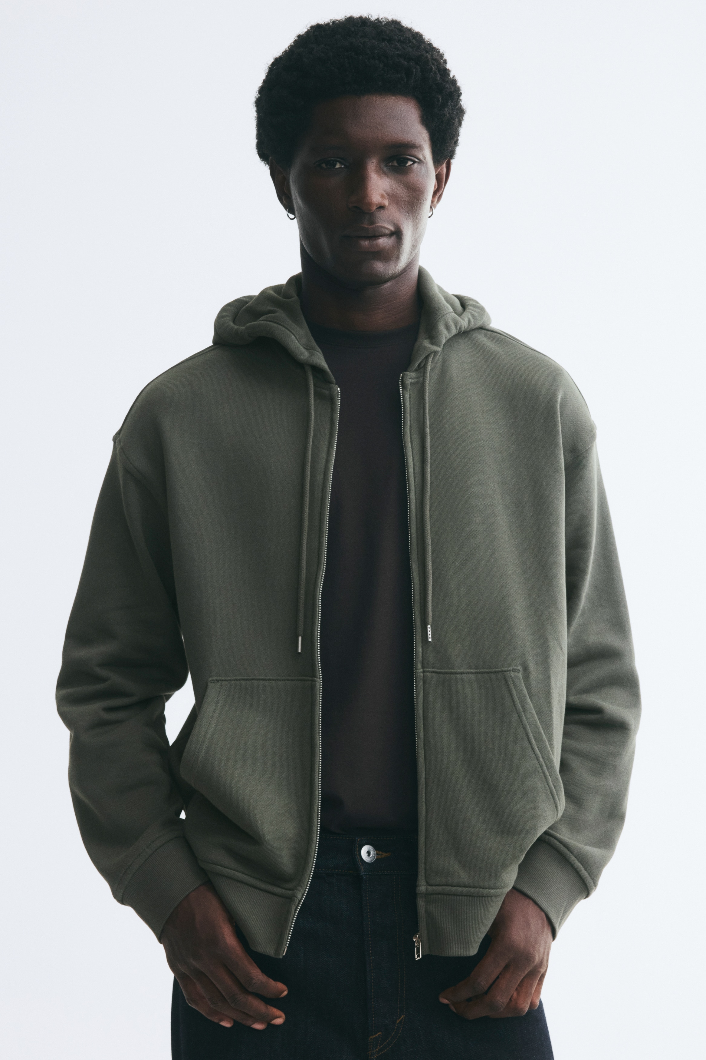 A dark green zip-up hoodie is shown from the front, featuring a relaxed fit with dropped shoulders and a drawstring hood. It has a full-length front zipper, a split kangaroo pocket, and ribbed finishes at the cuffs and hem.