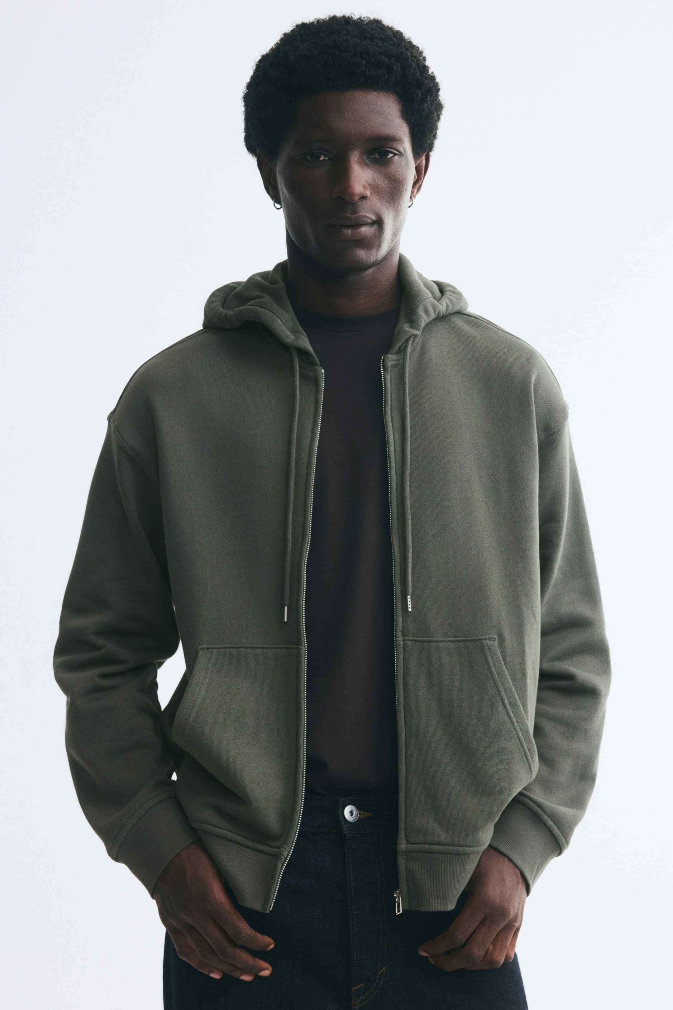 View larger image: A green zip-up hoodie is worn open by the model, featuring a drawstring hood, dropped shoulders, front pockets, and ribbed cuffs. The hoodie is layered over a dark T-shirt and paired with dark jeans, with the model standing with hands placed in the trouser pockets.