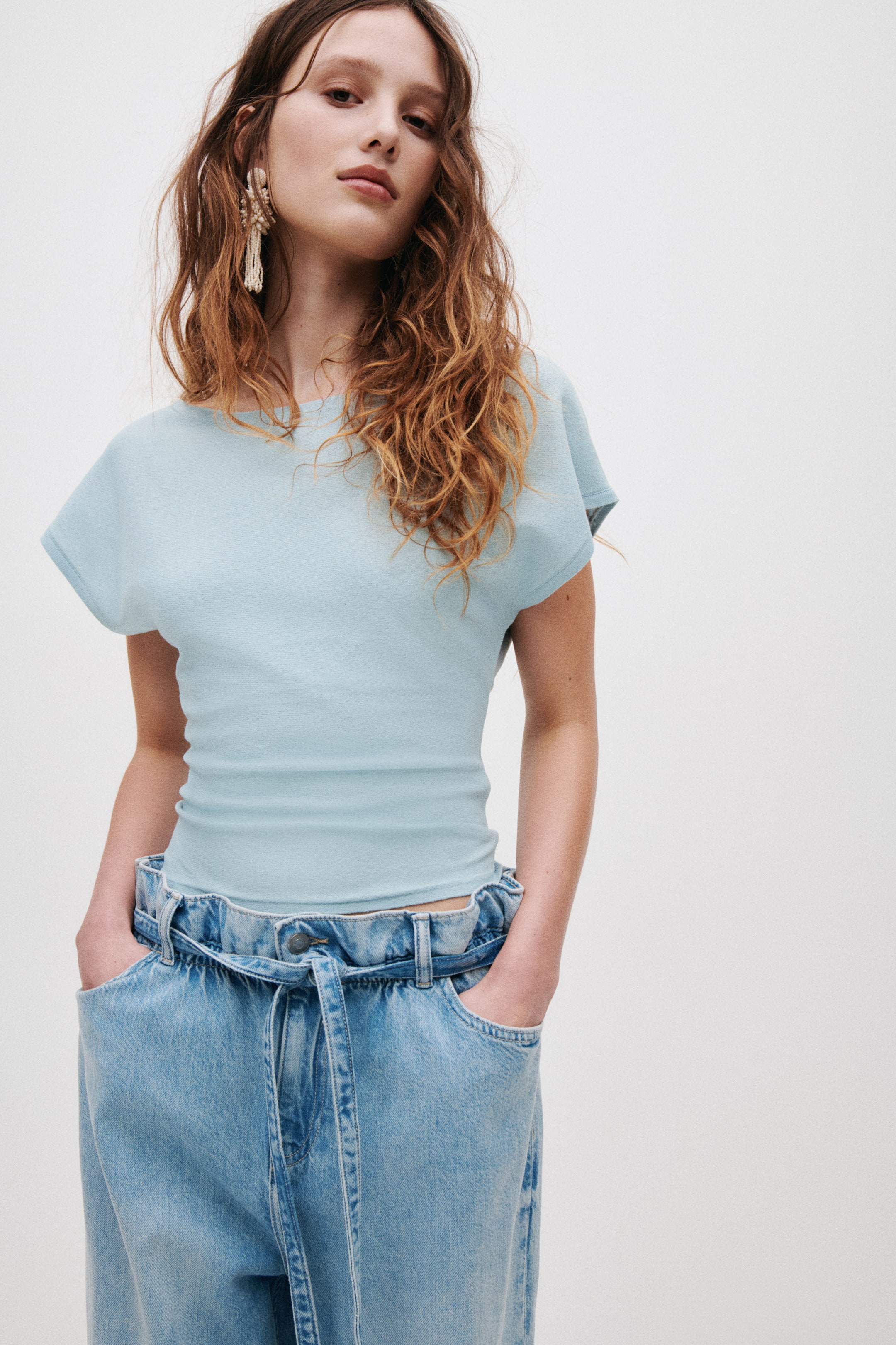 View larger image: The model is wearing a light blue, fine-knit top featuring a boat neckline, short sleeves, and a fitted silhouette. She's paired the top with high-waisted jeans.