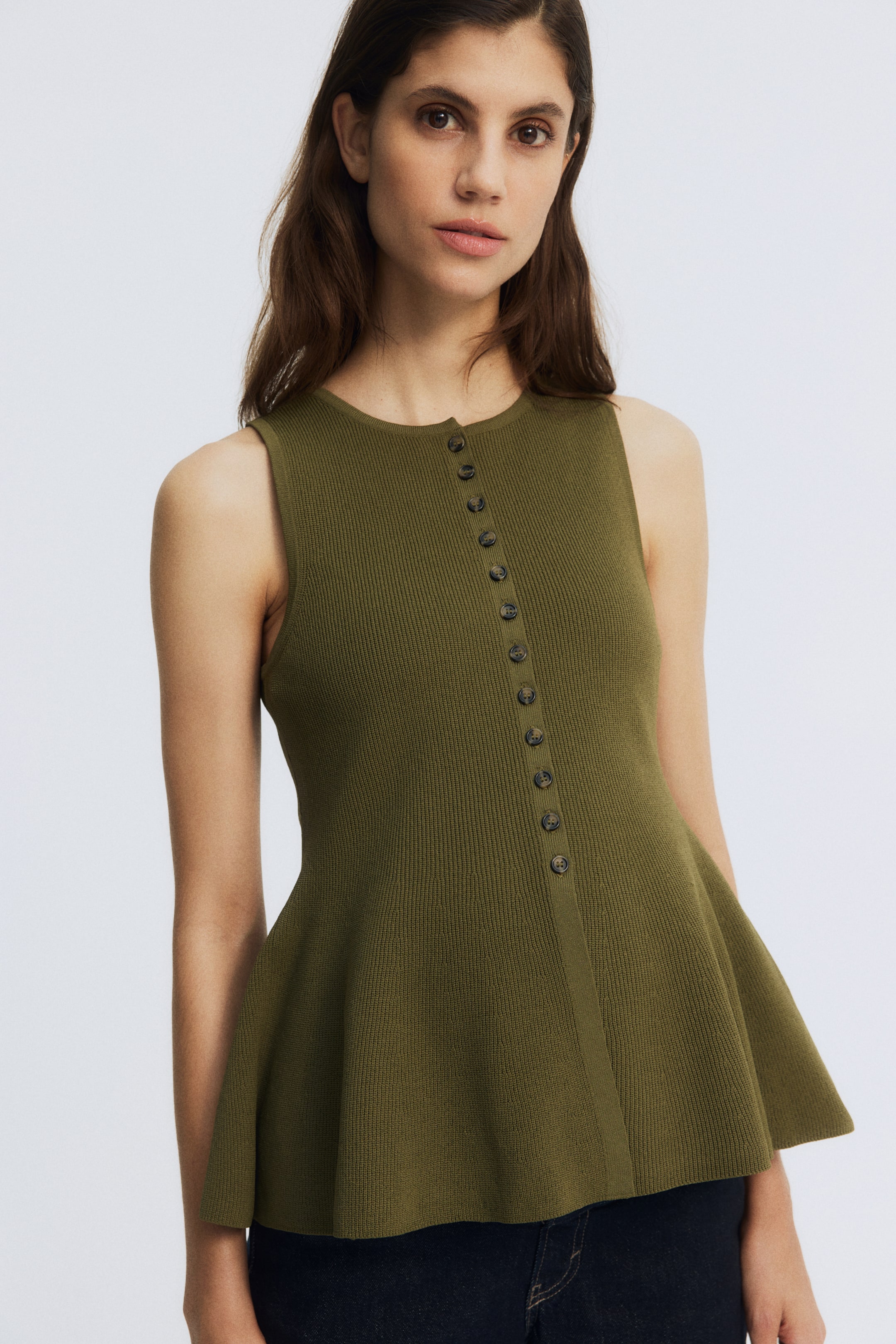 Ampliar la imagen: Un top sin mangas de punto acanalado en color verde oliva presenta un escote redondo y una hilera de botones completa con pequeños botones oscuros a lo largo del frente. El top cuenta con un dobladillo de peplum, que se ensancha desde la cintura y está ligeramente inclinado hacia la derecha, mostrando su ajuste y diseño.