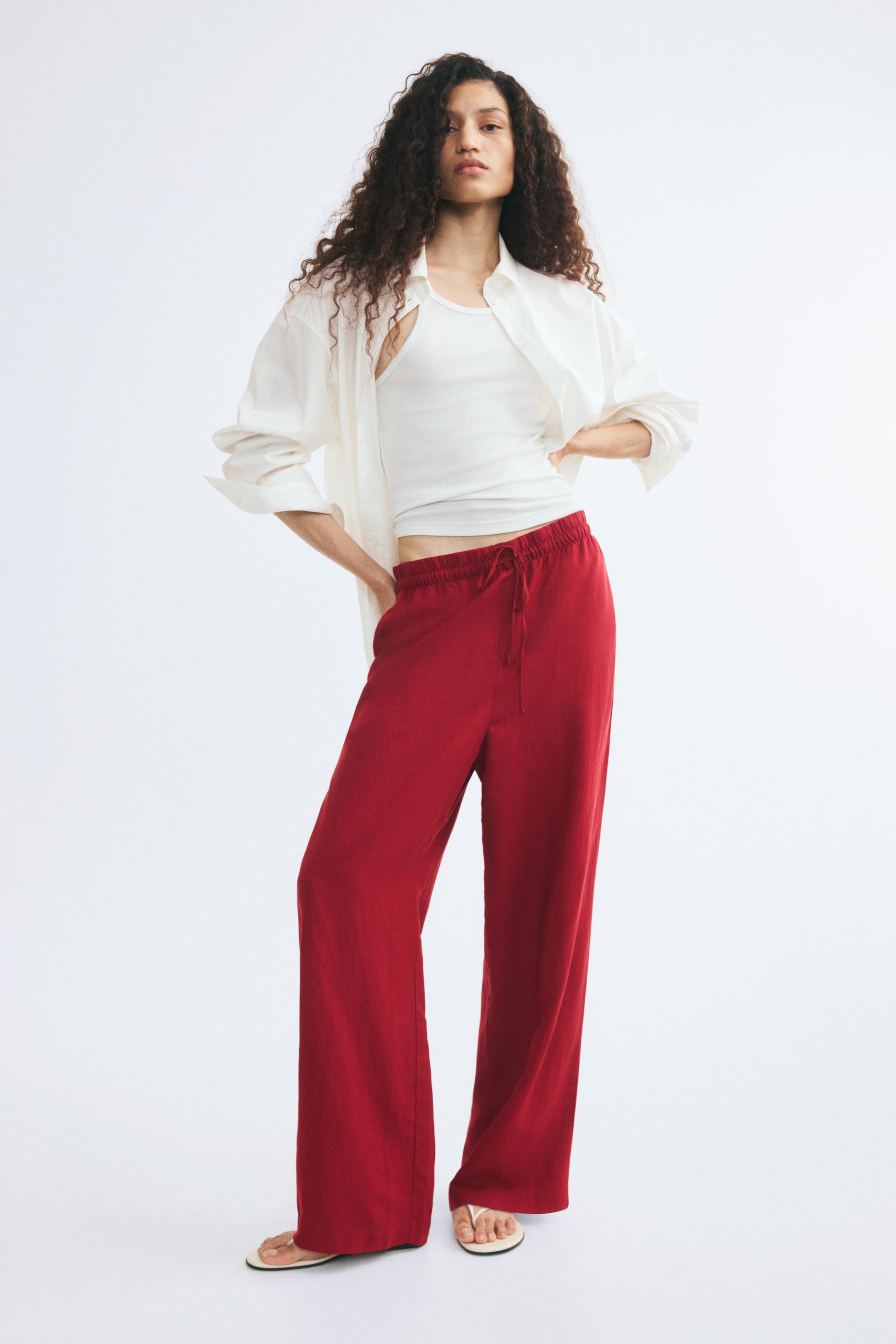 View larger image: The model is wearing red loose-fit trousers featuring a high, elasticated waist with a slim drawstring and straight legs. The trousers are styled with a white tank top and an open white long-sleeved shirt.