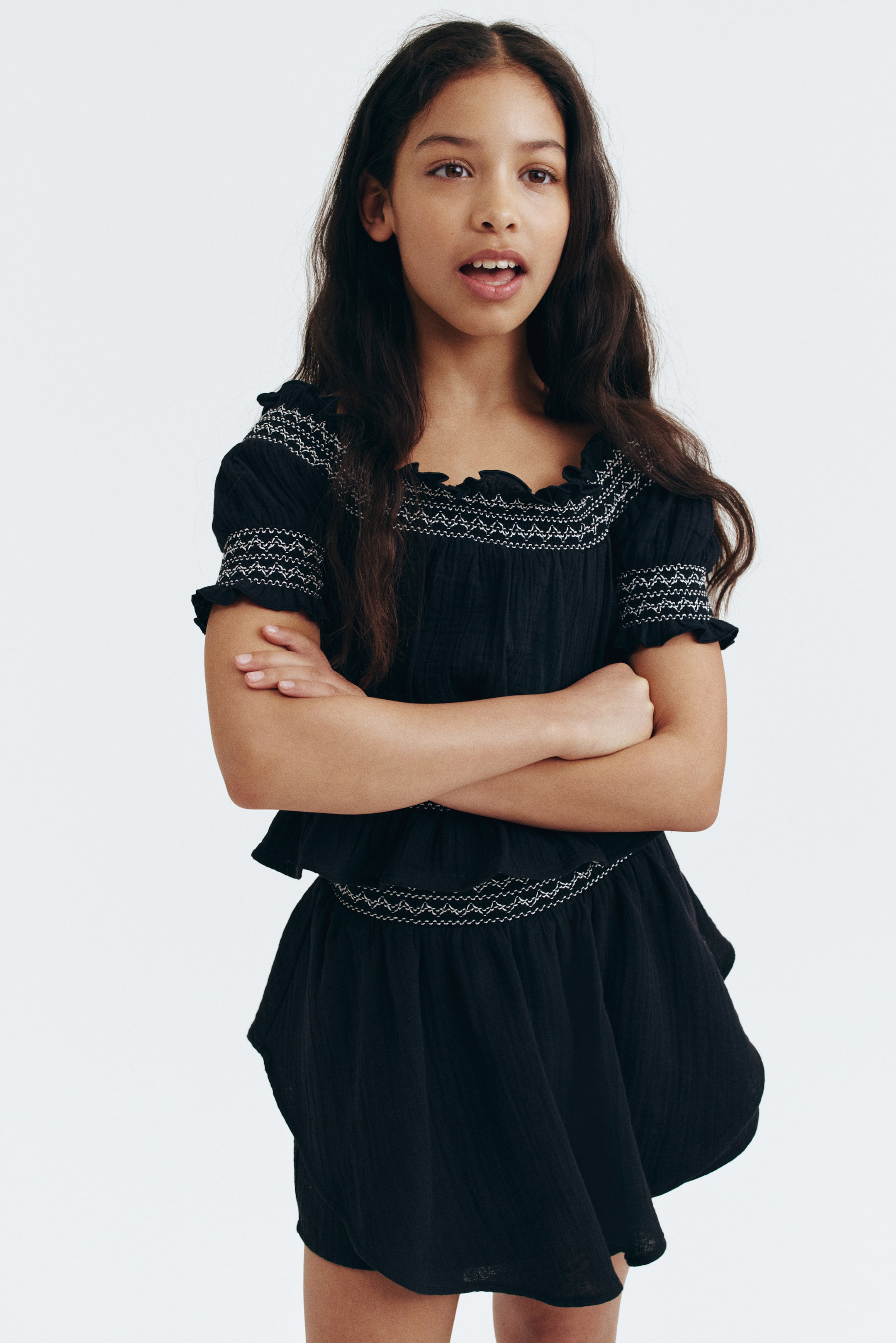 Grotere afbeelding bekijken: Een zwarte tweedelige outfit met een top en bijpassende shorts, beide voorzien van witte geborduurde smockdetails. De top heeft een vierkante halslijn met een gerafelde rand en korte pofmouwen met elastische gerafelde manchetten, terwijl de bijpassende shorts een gesmockte tailleband en een ontspannen, uitlopende pasvorm hebben.