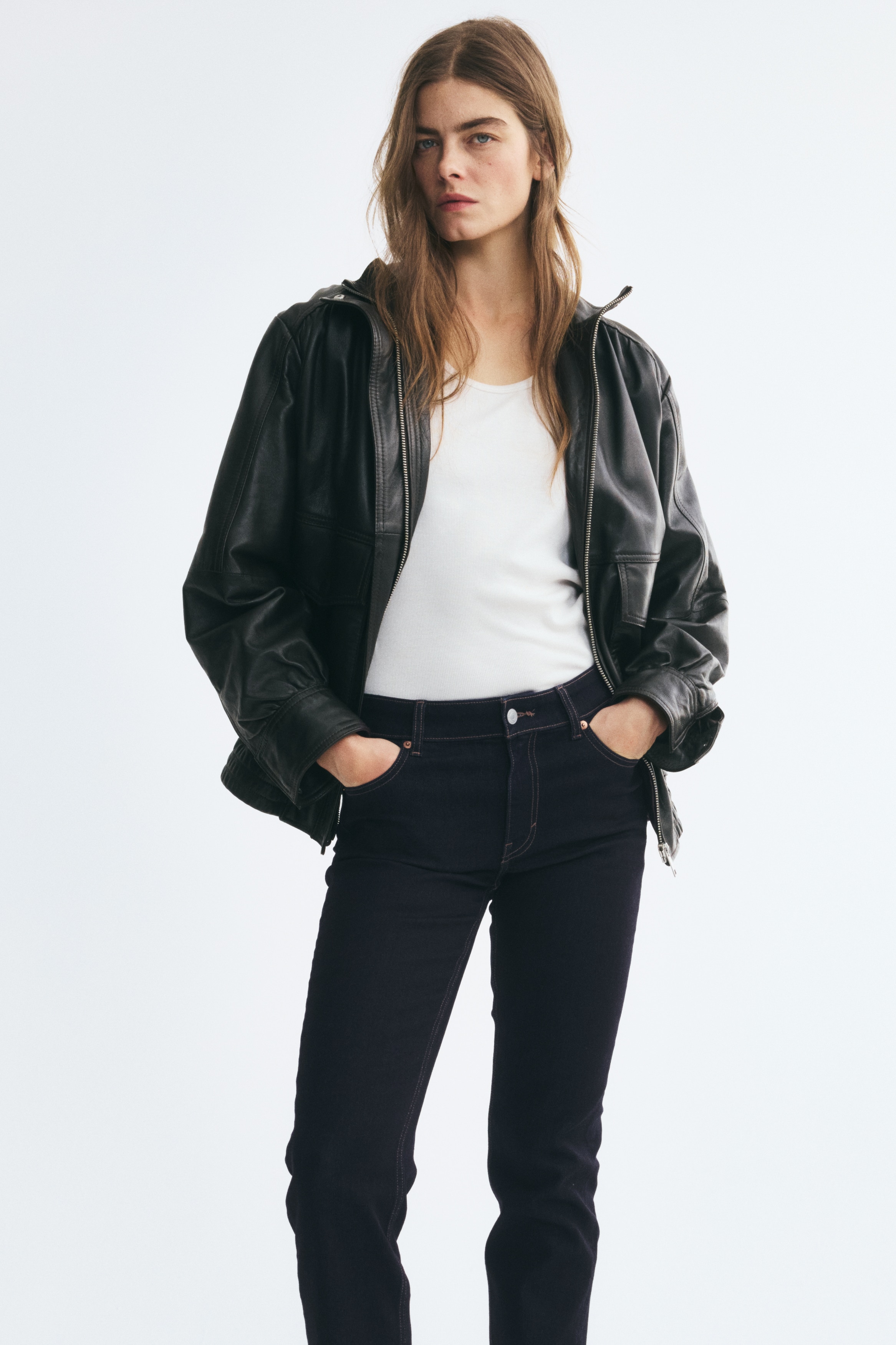 A pair of dark-wash indigo jeans is shown from the front, featuring a straight-leg silhouette with a classic five-pocket design. The jeans feature contrast orange stitching, copper rivets, and a silver-toned button fastening on the waistband.