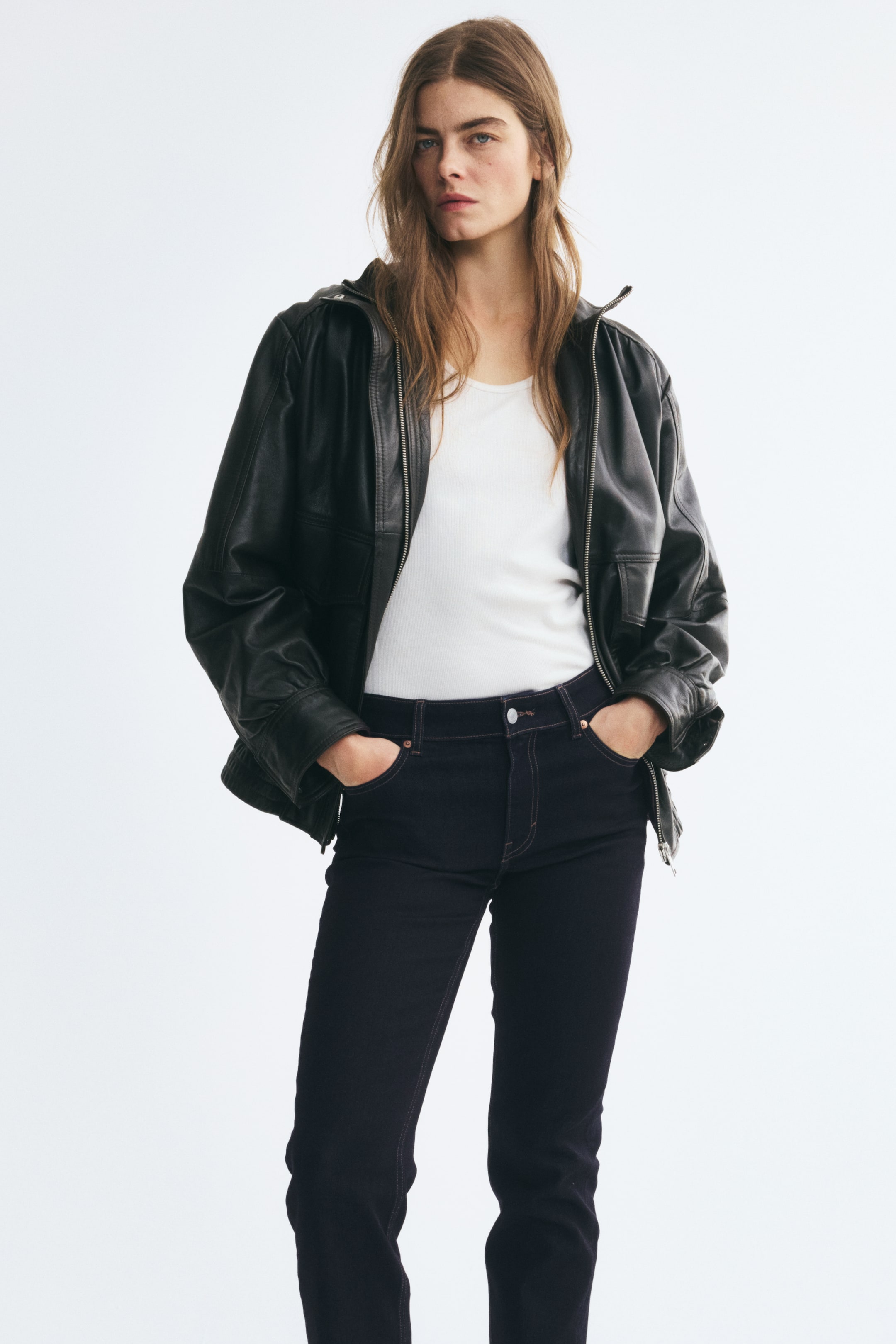 View larger image: The model is wearing dark indigo five-pocket denim jeans and is standing with their hands resting in the front pockets. The slim-fit, regular-waist jeans are styled with a tucked-in white T-shirt and an open black leather jacket.