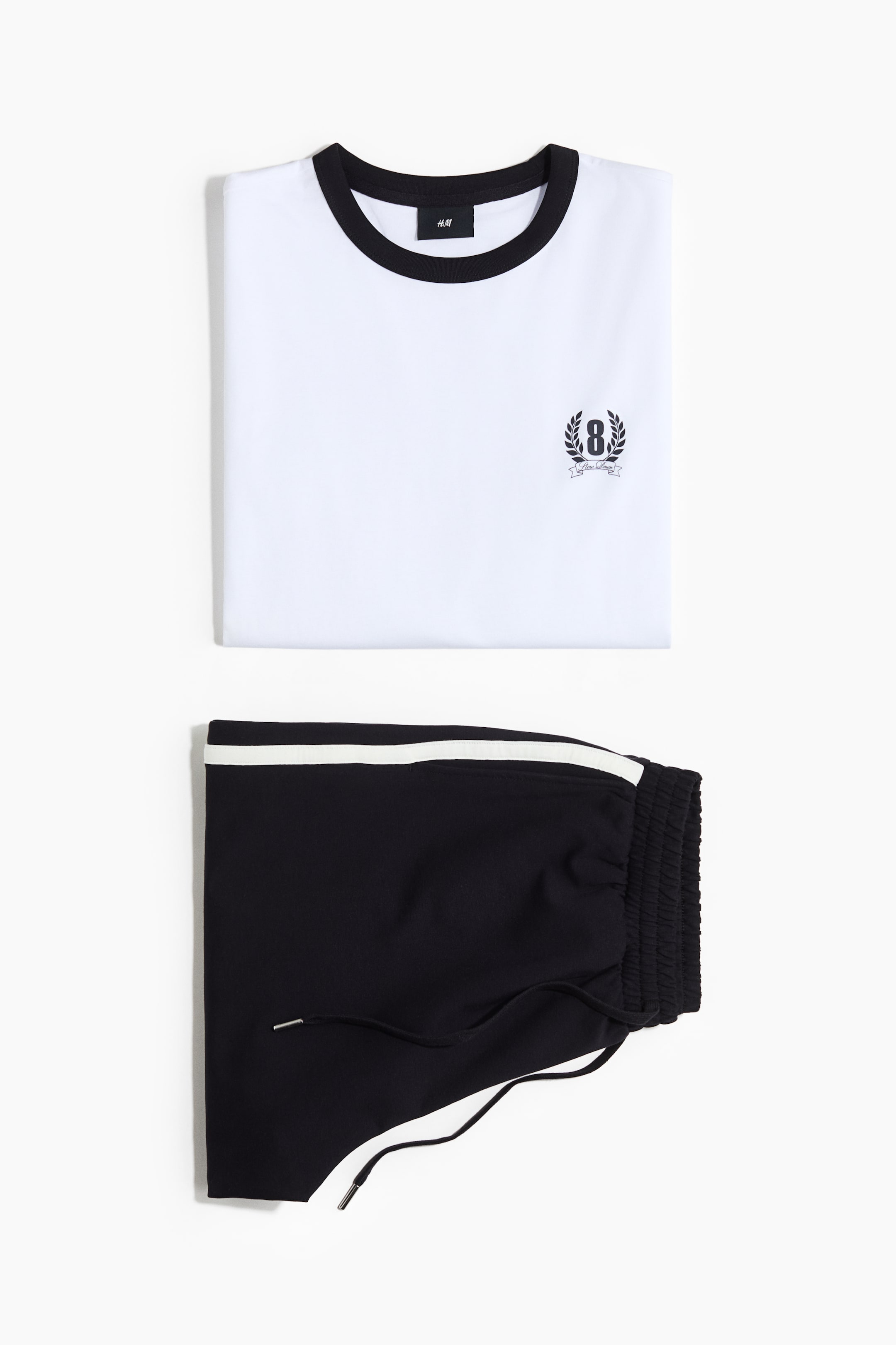 View larger image: A white t-shirt featuring a black-trimmed round neck and a small embroidered emblem showcasing the number '8' encircled by a laurel wreath is displayed on its front. Below it, black shorts are highlighted by a white stripe along the side, an elasticated waistband, and a black drawstring with silver tips.