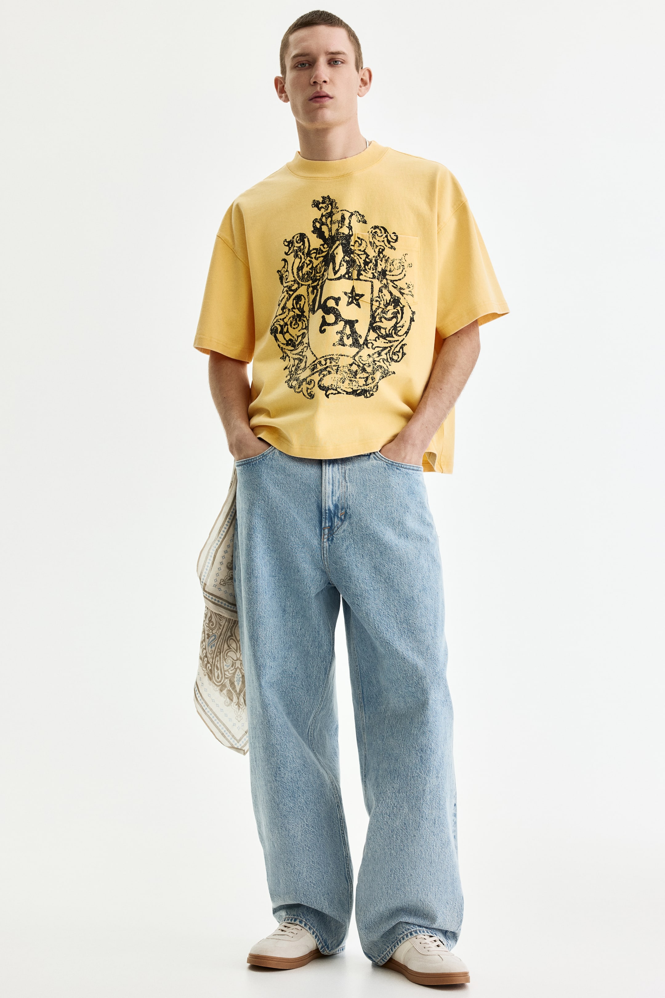 View larger image: A yellow oversized short-sleeve T-shirt displays a large black crest graphic on the front, paired with light blue wide-leg jeans. A patterned scarf hangs from the left front pocket of the jeans, and cream low-top sneakers with light brown soles are worn.