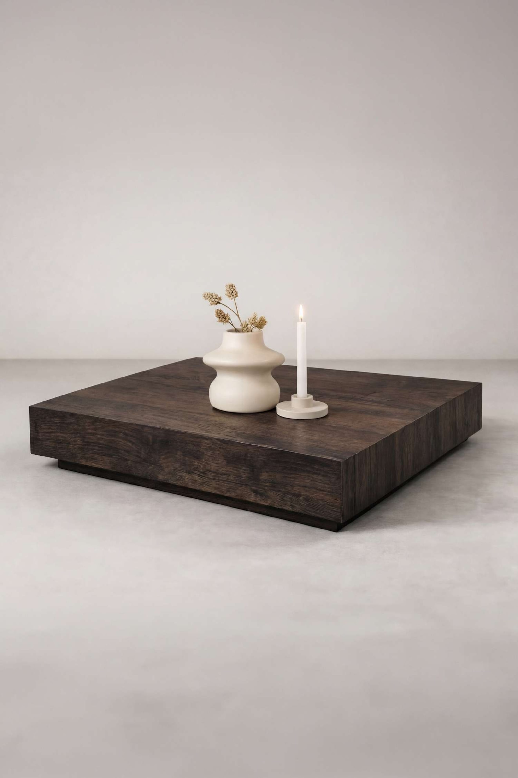 Größeres Bild ansehen: Ein quadratischer Couchtisch aus dunklem Holz zeichnet sich durch ein niedriges Profil und sichtbare Holzmaserung auf der Oberseite und den Seiten aus. Eine cremefarbene Vase mit getrockneten Pflanzen und eine brennende weiße Kerze in einem Halter stehen auf seiner Oberfläche.