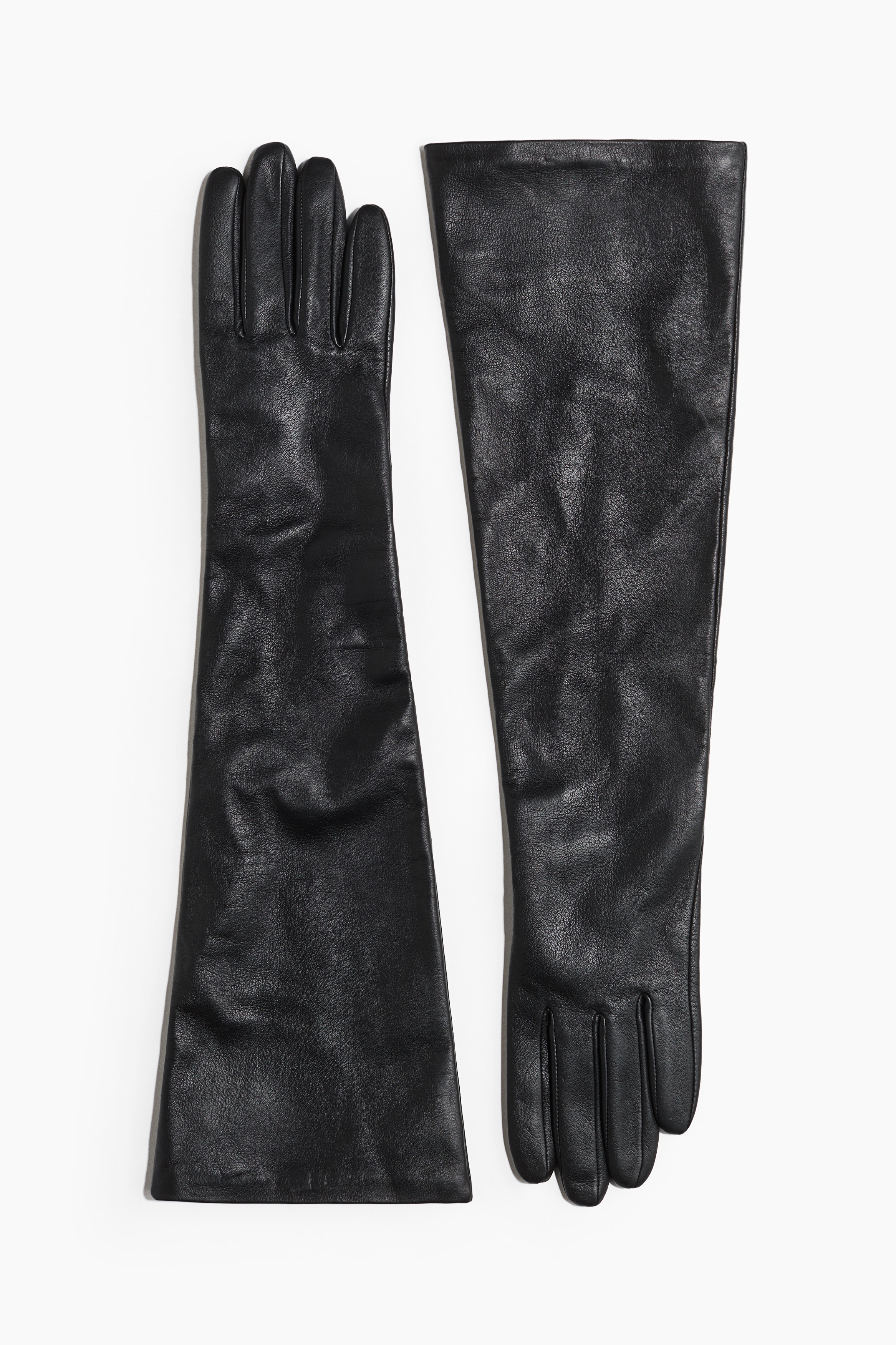Long black leather gloves featuring a wide, slouchy design that extend past the elbow are being worn by the model. The gloves are layered over a long-sleeved black top, with the modelâs arms crossed in front of their waist.