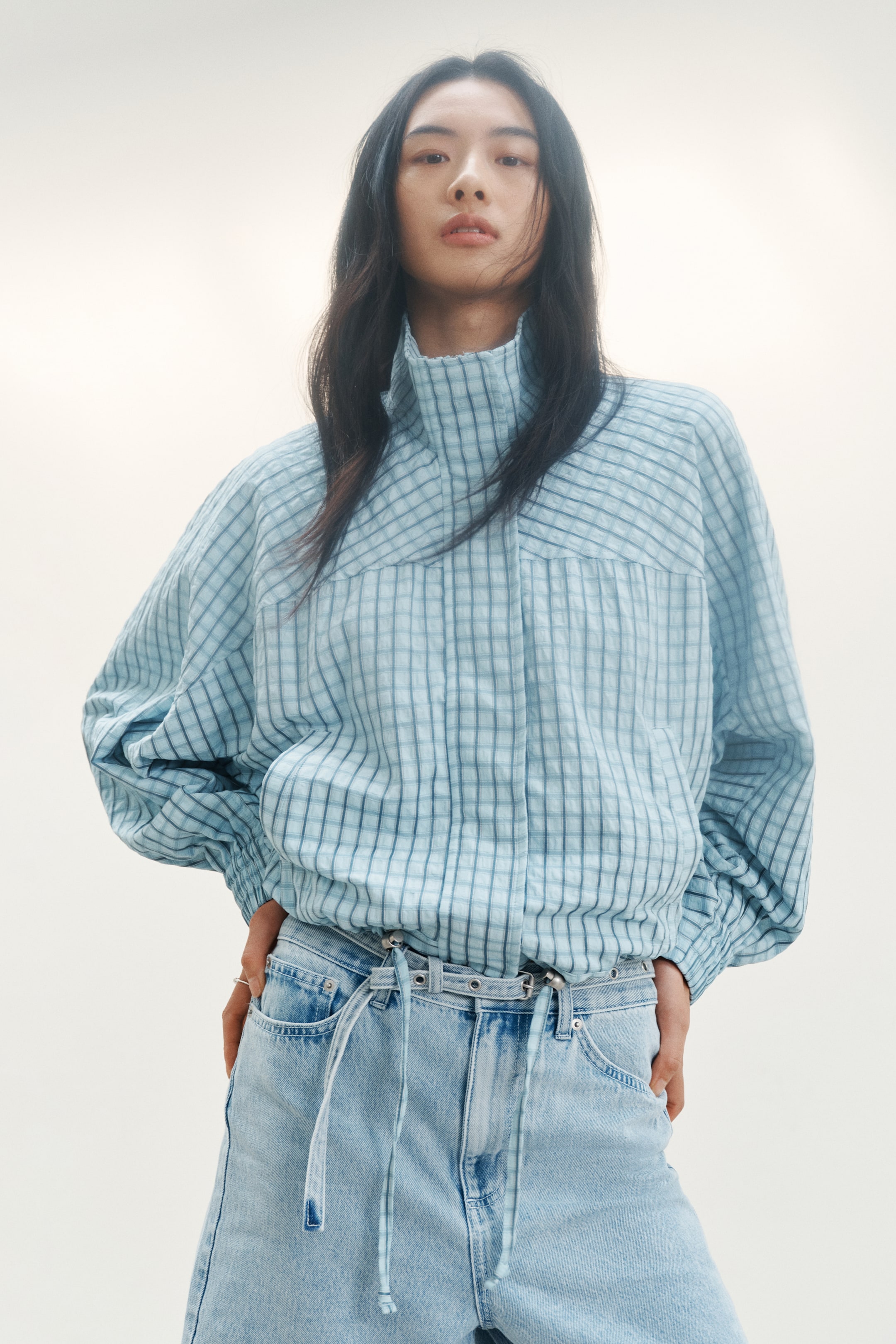 View larger image: A light blue jacket crafted from crinkled check fabric showcases a high stand-up collar and a prominent upper chest yoke. Designed with voluminous long sleeves that end in elasticized cuffs and an elasticized hem, it offers a blouson fit. The jacket is styled facing forward with the hem resting just above a visible belt, perfect for a chic and contemporary look.