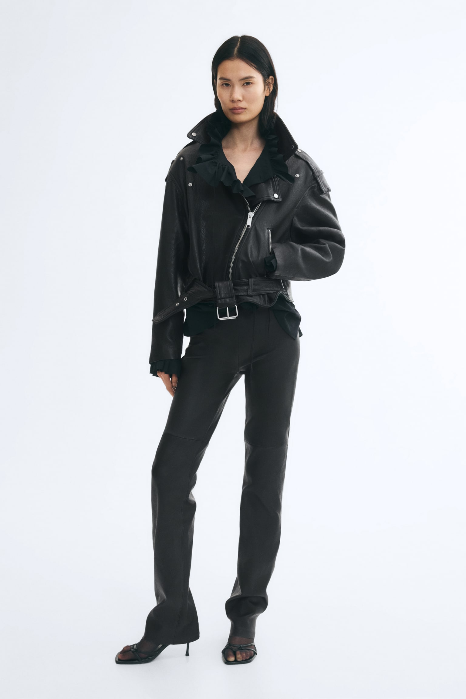 En sort lÃ¦derjakke med krÃ¸llet look, med asymmetrisk lynlÃ¥s foran, brede revers og et justerbart bÃ¦lte i kanten, bÃ¦res af modellen. Jakken styles over en sort skjorte med flÃ¦ser og matchende smalle bukser, mens modellen hviler den ene hÃ¥nd i sidelommen. - 1