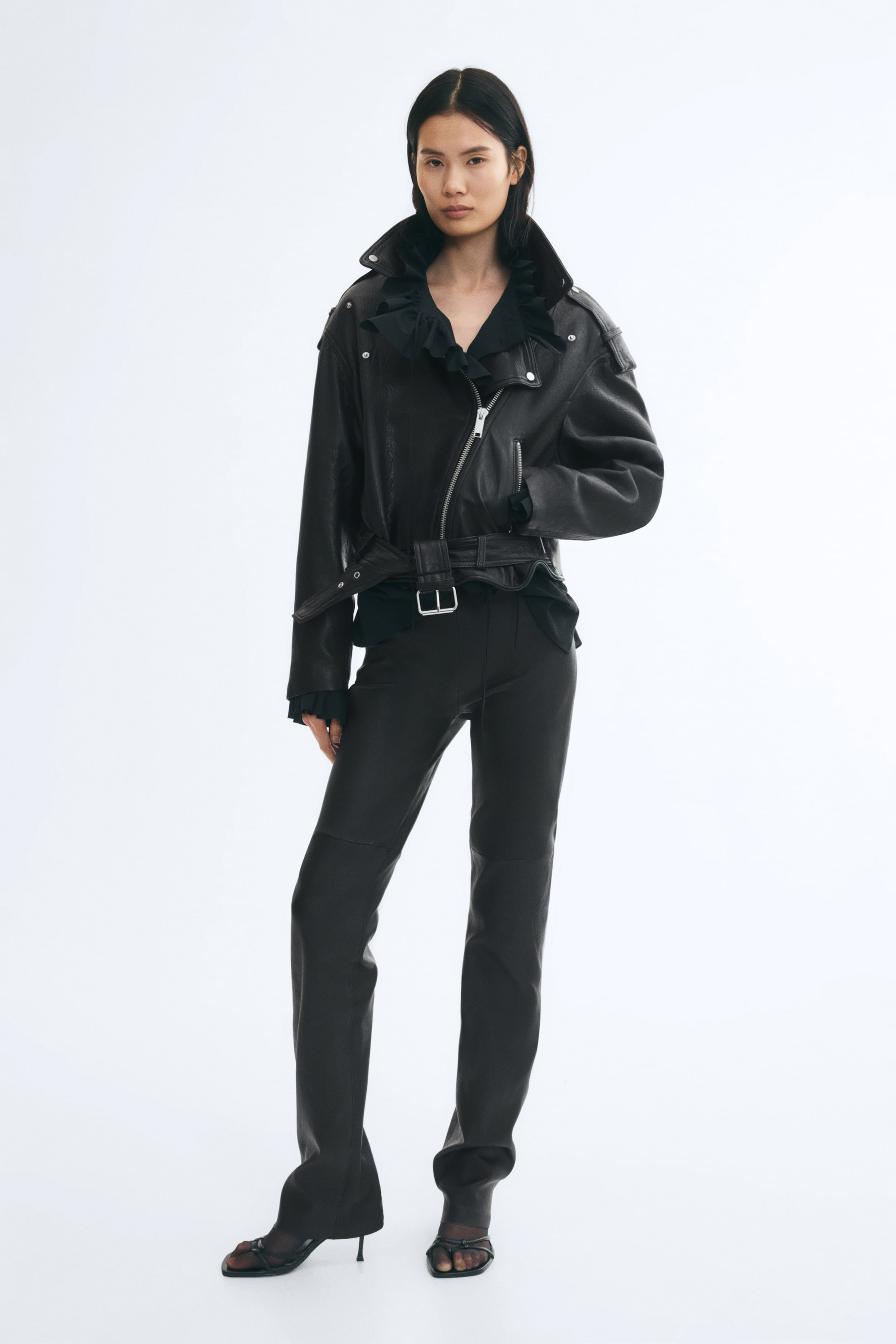 Se større billede: En sort læderjakke med krøllet look, med asymmetrisk lynlås foran, brede revers og et justerbart bælte i kanten, bæres af modellen. Jakken styles over en sort skjorte med flæser og matchende smalle bukser, mens modellen hviler den ene hånd i sidelommen.