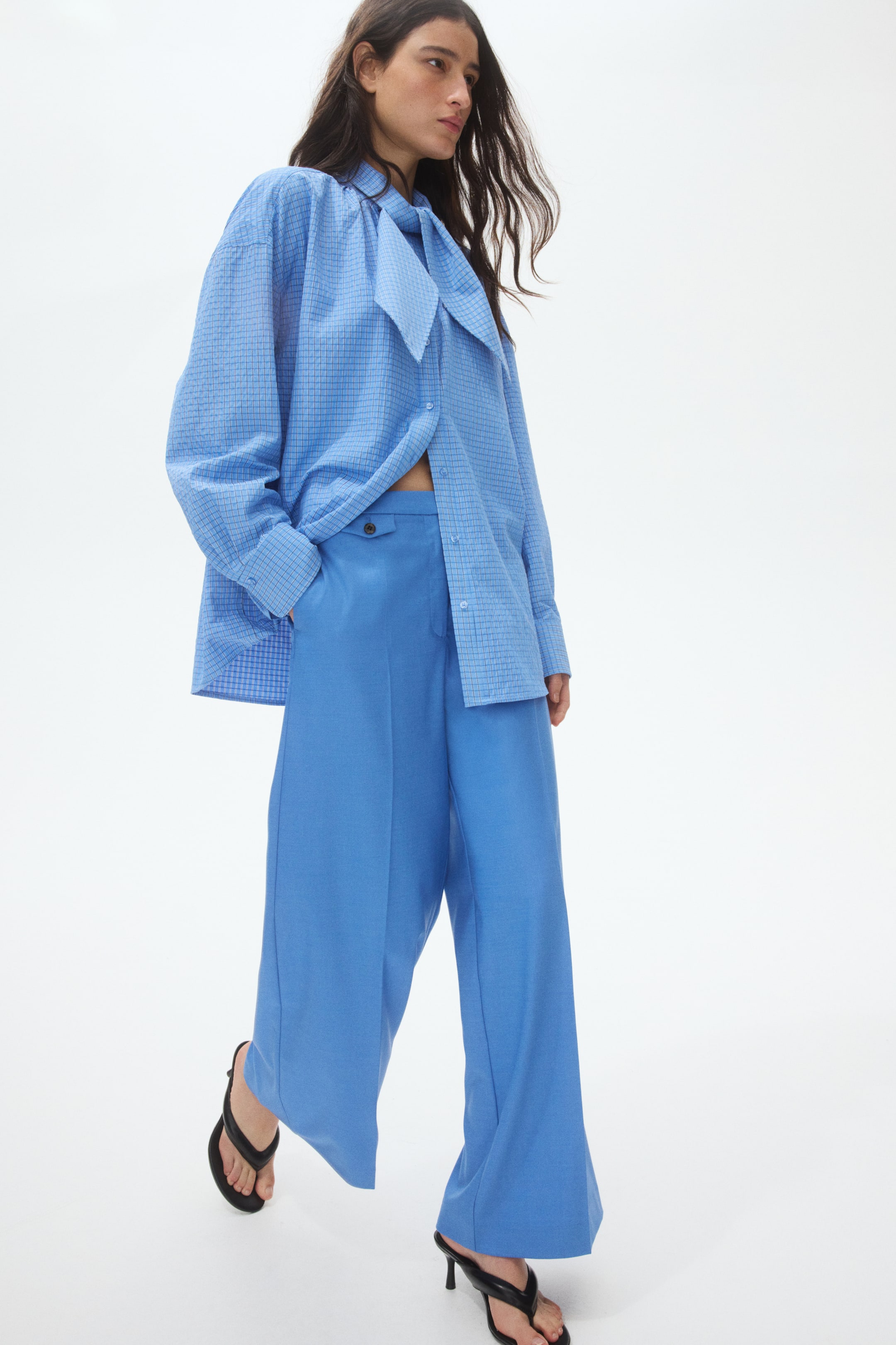 View larger image: An oversized light blue checkered shirt with a prominent pussy-bow necktie detail is styled with solid blue wide-leg trousers. The shirt is partially unbuttoned, while the trousers feature a button and tab closure on the left side.