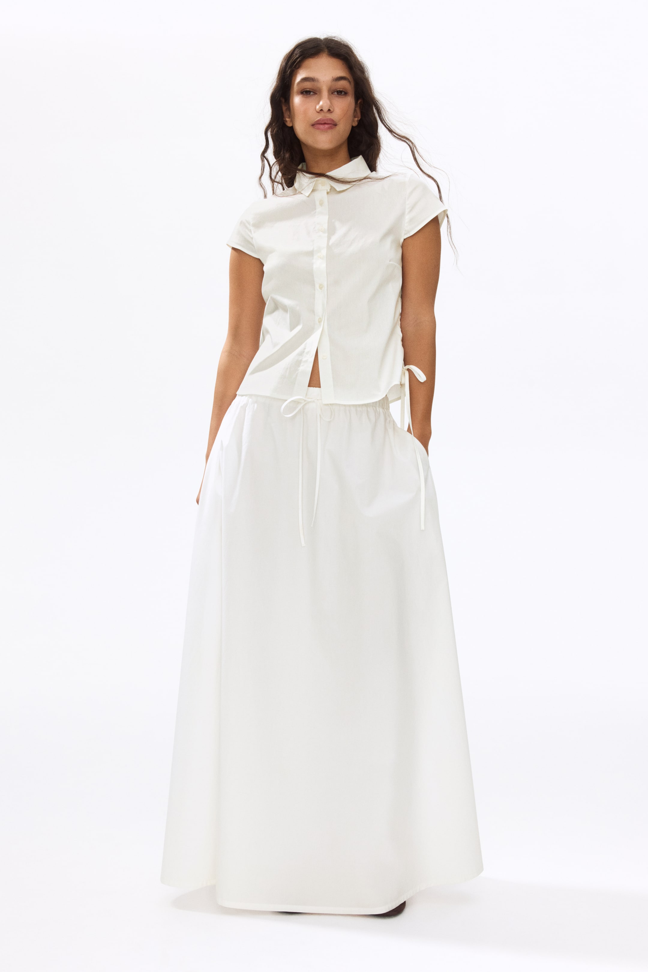 View larger image: A white two-piece outfit is presented from the front. The short-sleeved button-up shirt features a subtle vertical texture, a pointed collar, short sleeves, a partially unbuttoned front, and a cropped hem with side ties. The matching long maxi skirt showcases an elasticated drawstring waistband, side pockets, and a flowy silhouette.