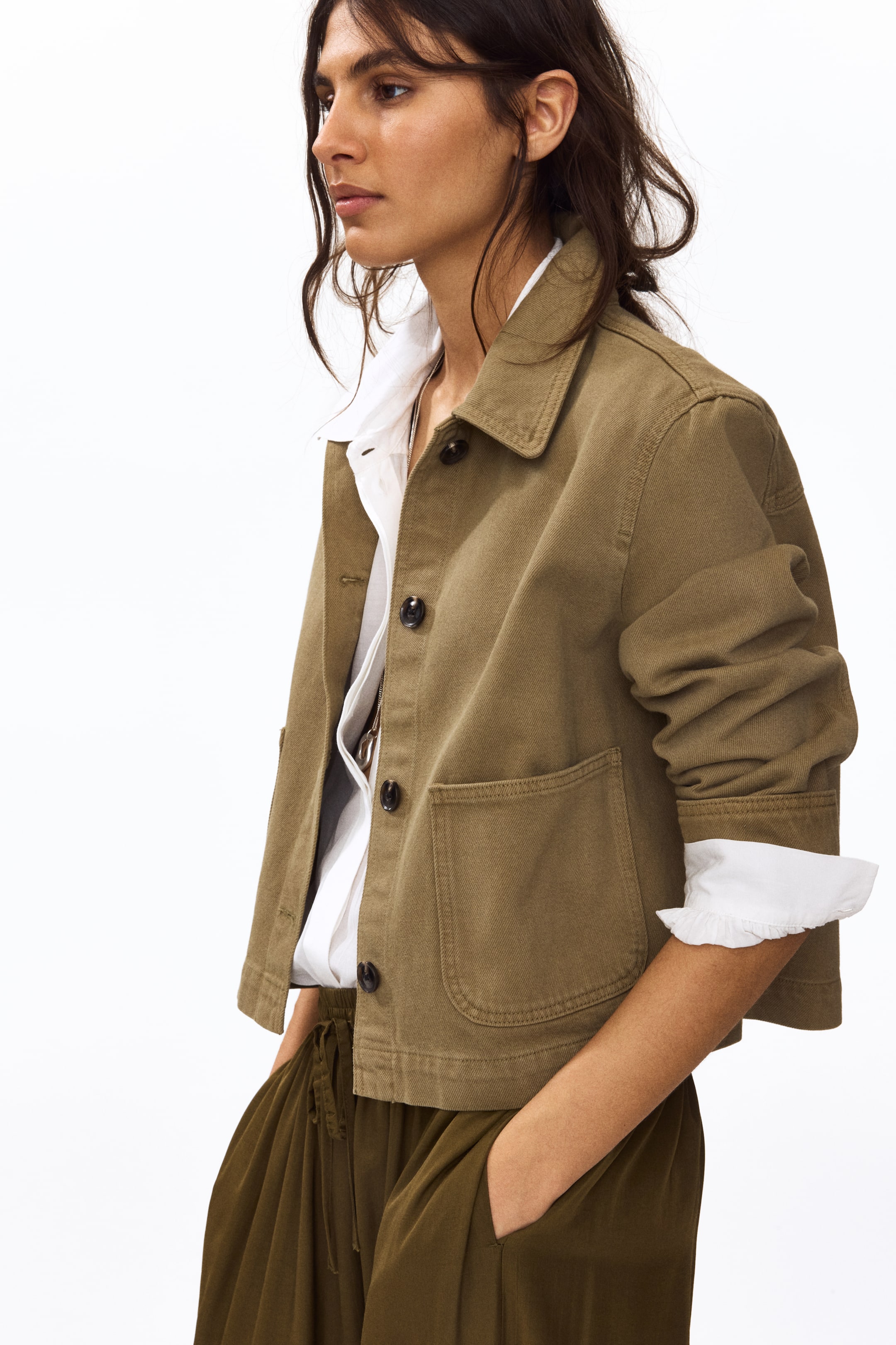 Visa större bild: En olivgrön kort jacka med spetsig krage, full knäppning framtill med mörka knappar och två framfickor, bärs över en vit skjorta med uppvikta manschetter över jackans ärmar.