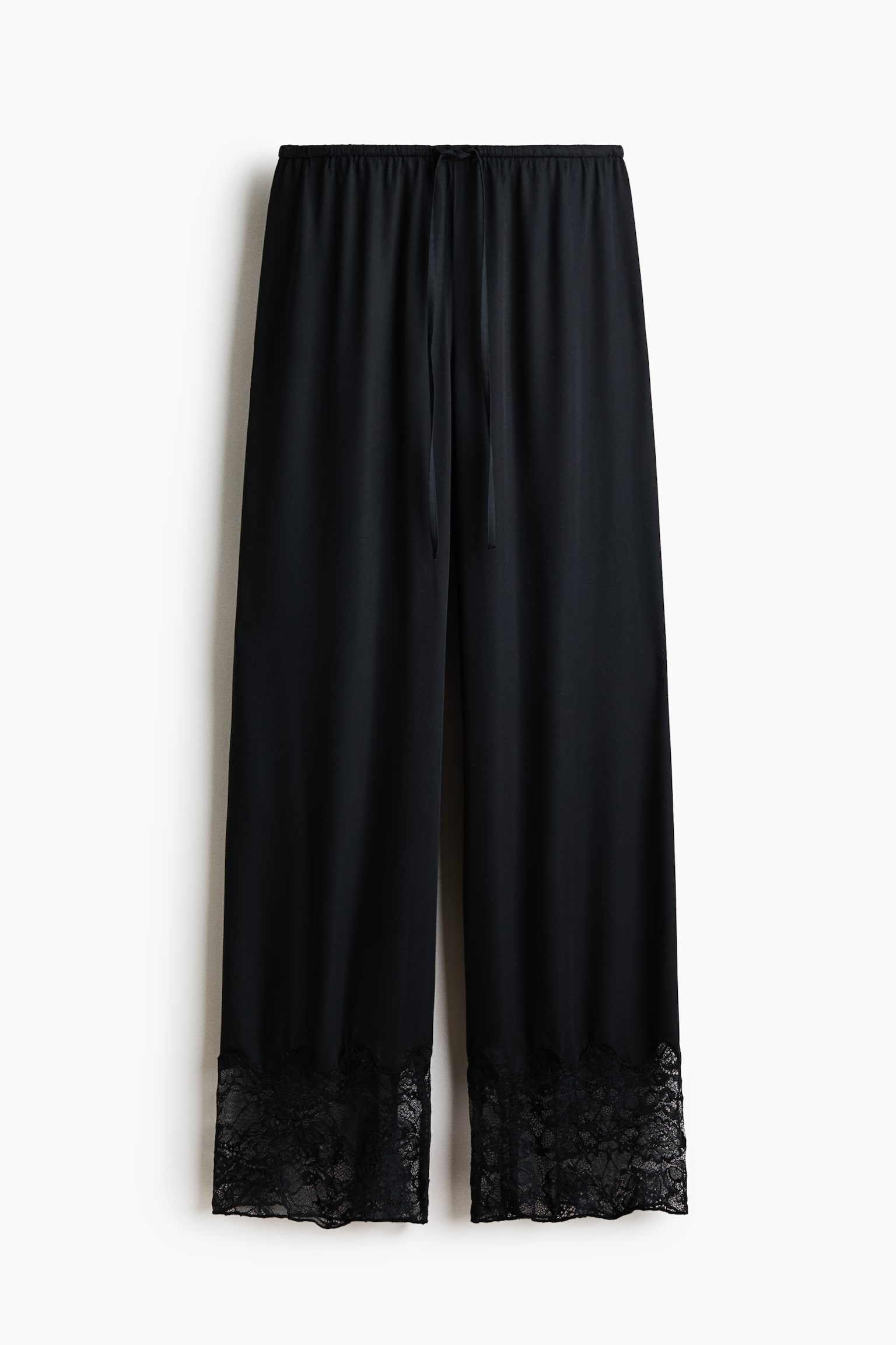 Black pyjama bottoms are hanging front-on, featuring an elasticated drawstring waist. They are showcasing a softly draping viscose satin fabric and are displaying wide black lace trim at the hems. - 1