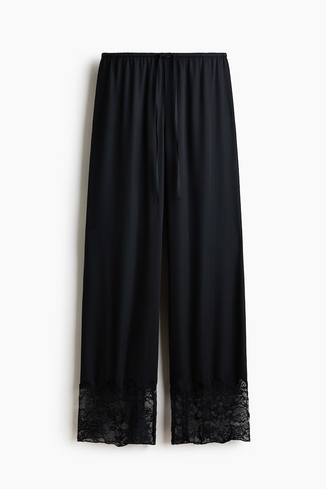 Black pyjama bottoms are hanging front-on, featuring an elasticated drawstring waist. They showcase a softly draping viscose satin fabric and display wide black lace trim at the hems. - 1